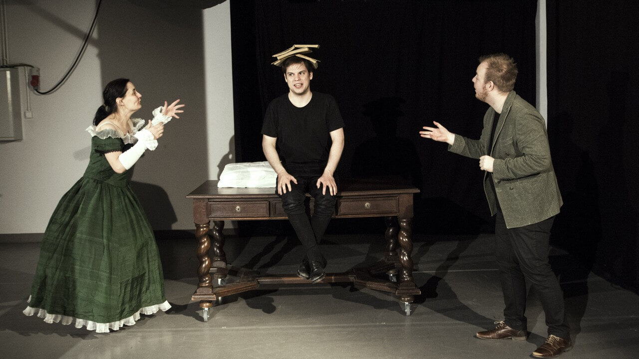 Zdjęcie ze spektaklu Bajka o księciu Pipo. Przedstawia trójke aktorów. Mężczyzna siedzi centralnie na stole z 3 ksiażkami na głowie. Aktorka po lewej stronie w obszernej zielonej sukni. Z prawej aktor w szarej marynarce.