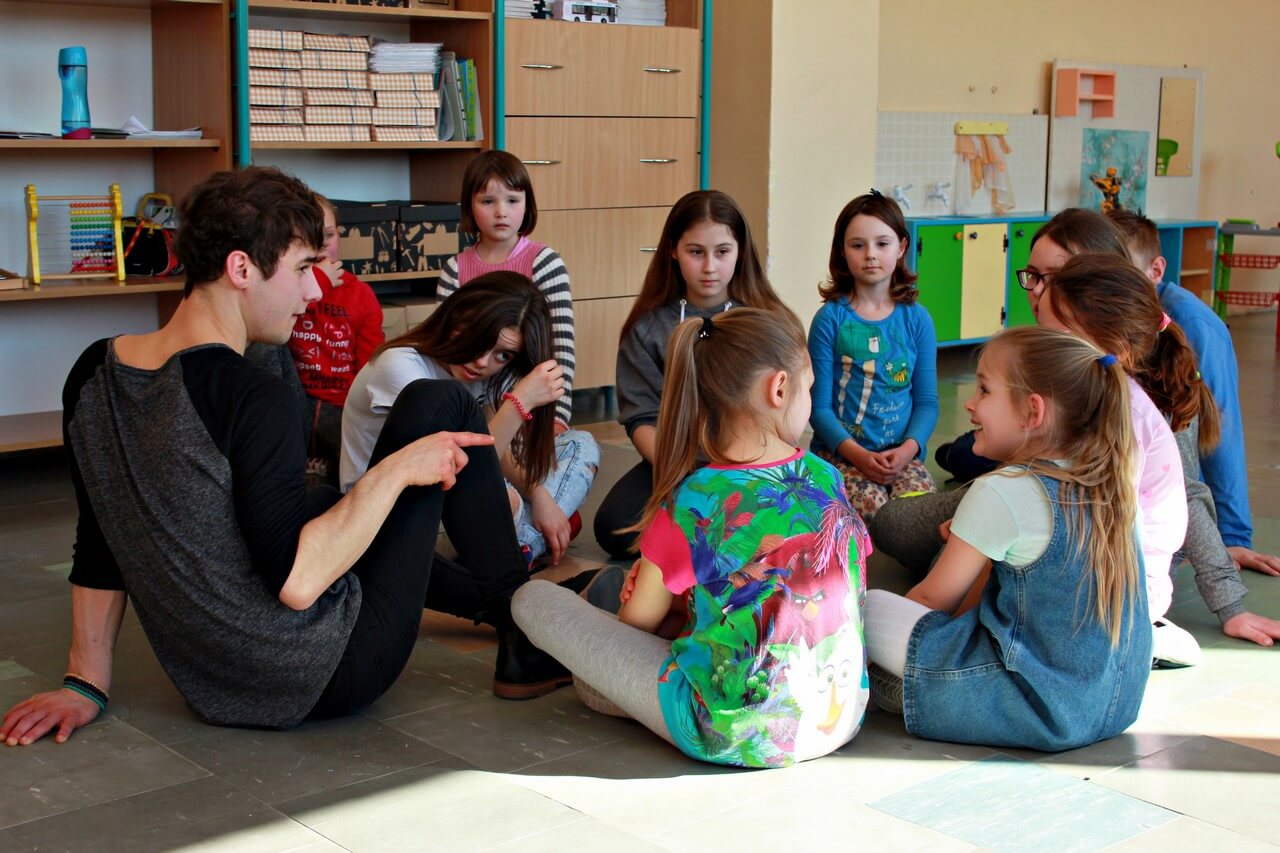 Na zdjęciu grupa dzieci siedzących w okregu na podłodze i słuchających instruktora.
