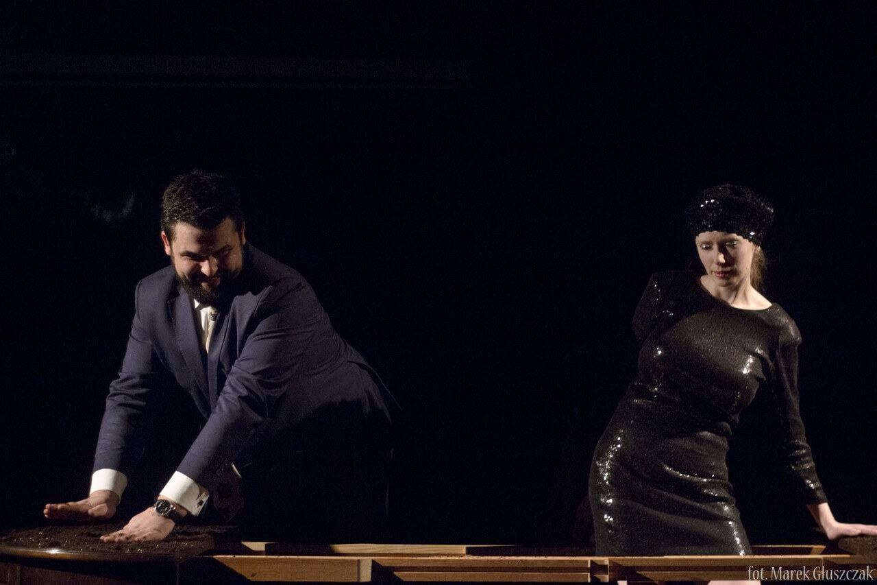 Zdjęcie ze spektaklu Burmistrz. Pokazuje dwoje aktorów przy stole. aktor w granatowym garniturze po lewo, aktorka w czarnej, ślniącej sukience po prawo.