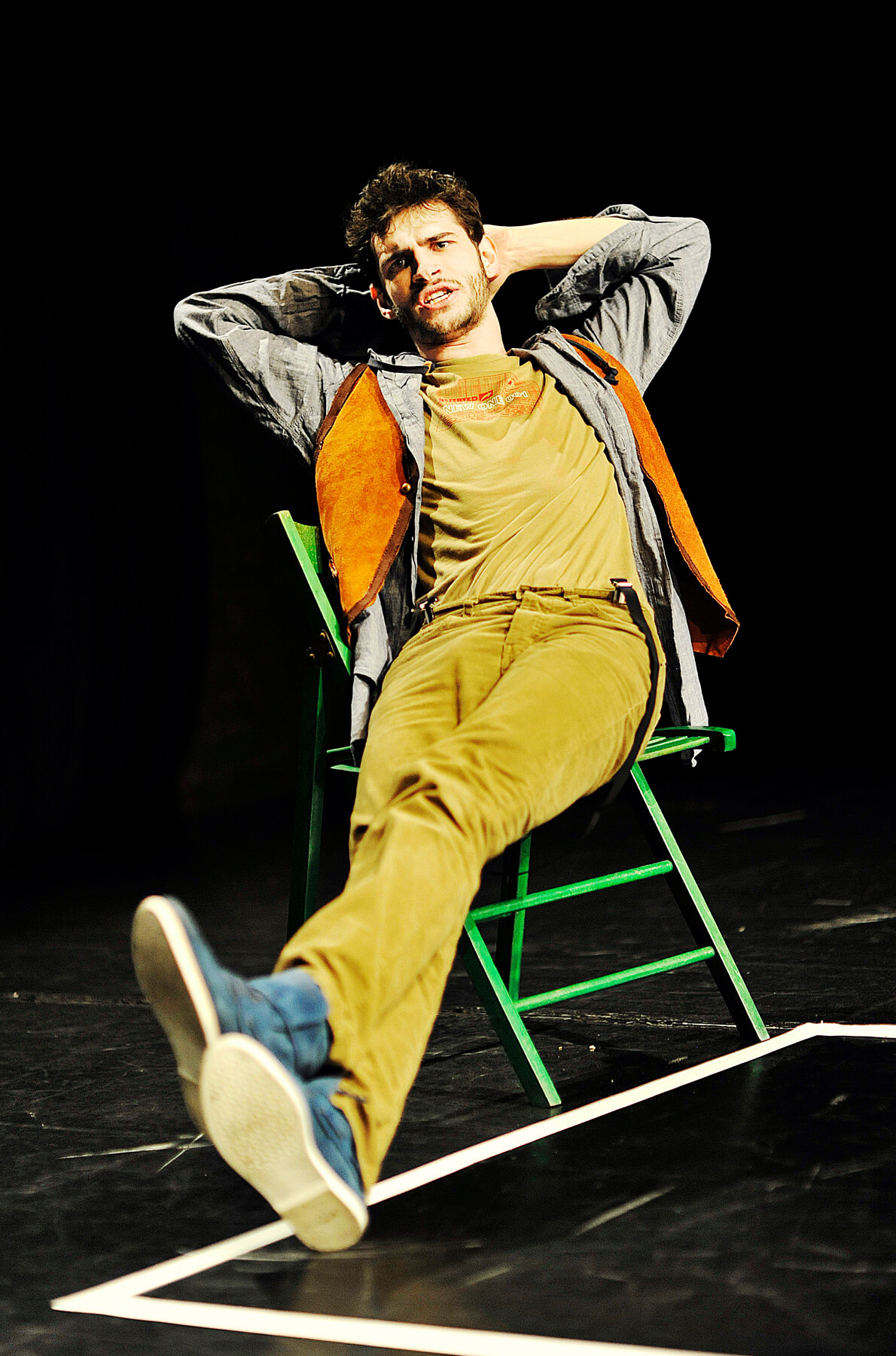 Zdjęcie do spektaklu Ścianananaświat. Aktor siedzi na zielonym krześle, z nogami wyciągnietym przed siebie i z rękami założonymi za głowę. Ubrany w niebieskie buty, zółte sztruksy, szara koszule i pomarańczową kamizelkę.