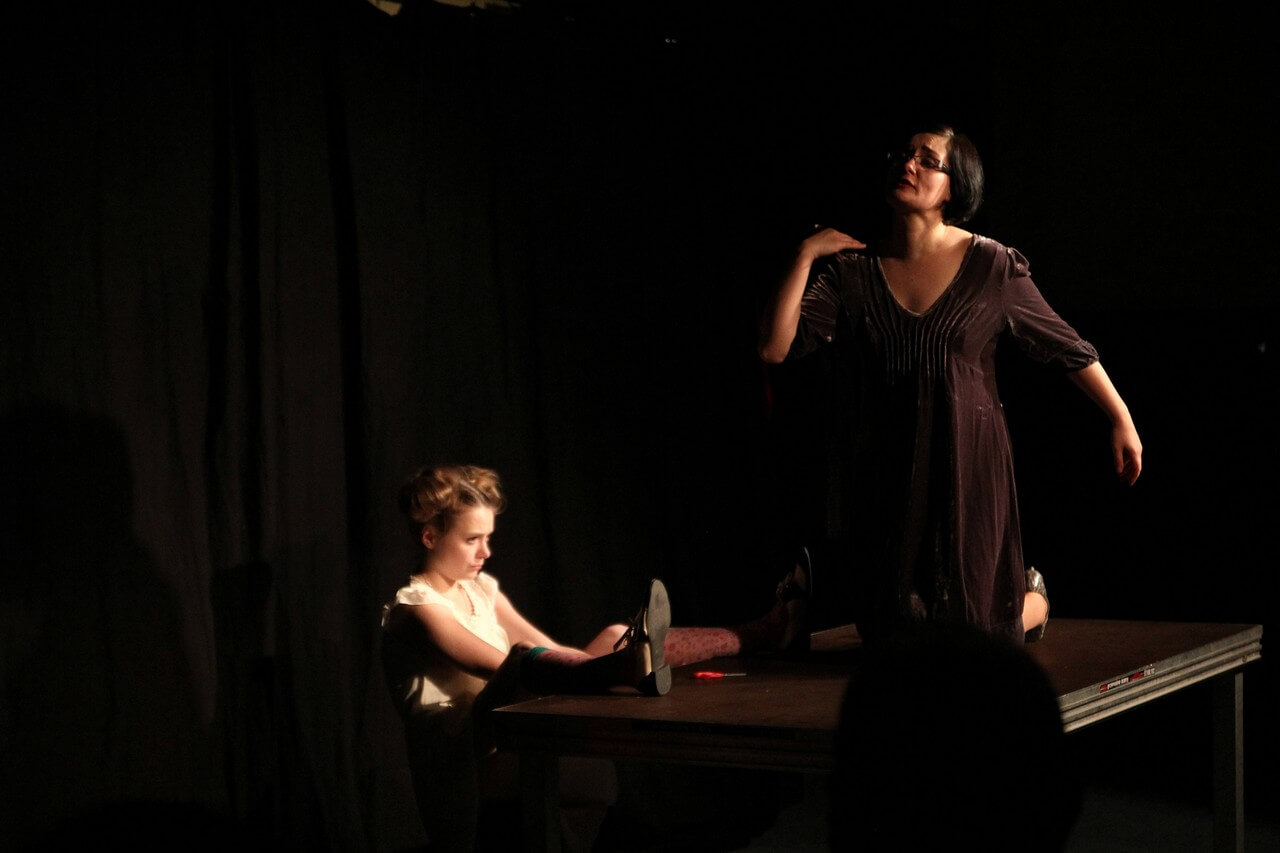 Zdjecie do spektaklu Kawa, chleb i ser. Aktorka w czarnej sukience klęczy na stole. Aktorka w białej sukience siedzi z lewej strony stolu z nogami zarzuconymi na blat.