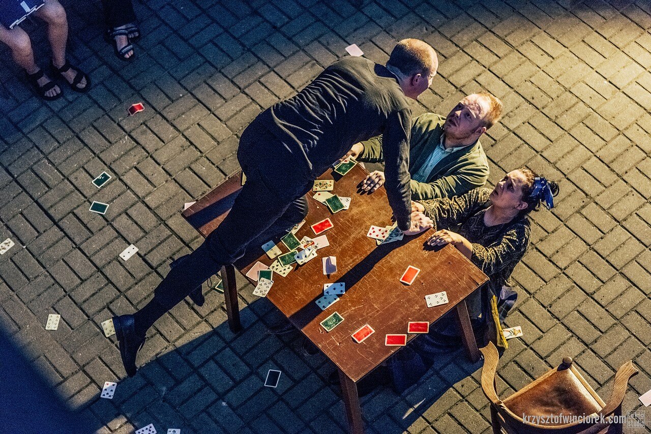 Aktor klęczy na stole i pochyla się w kierunku dwójki aktorów klęczących za stołem. Wszedzie leżą rozsypane karty do gry.