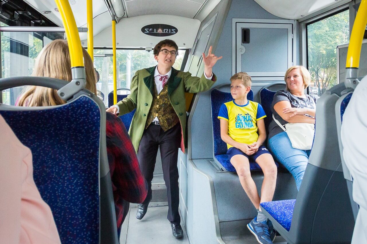 Zdjęcie w autobusie, elegancko ubrany młody mężczyzna w okularach wykrzykuje coś stojąc między fotelami, obok widać innych pasażerów, po lewej tył postaci w czerwonej koszuli w kratę, po prawej siedzących młodego chłopca w stroju Arki Gdynia i dorosłą kobietę w krótkich blond włosach