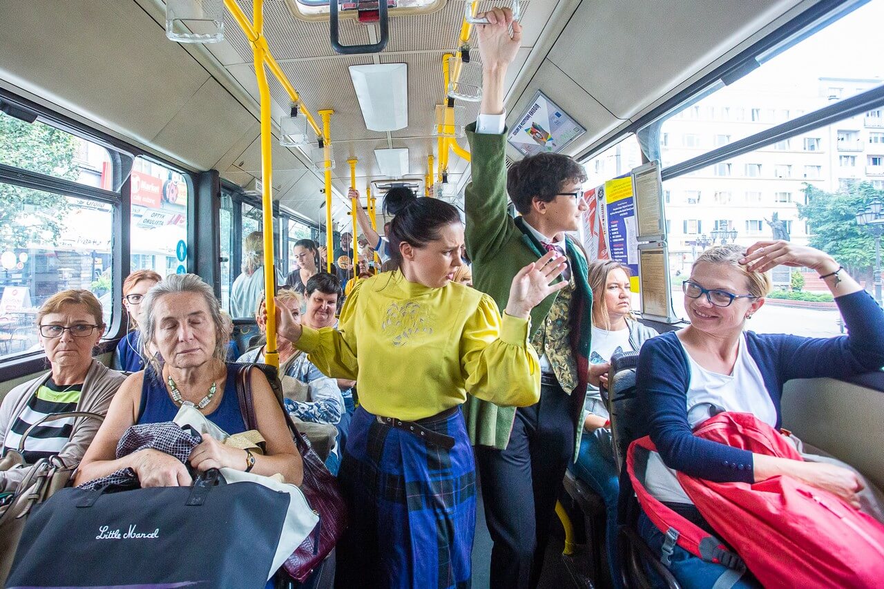 wnętrze autobusu, na środku dwie postacie, po lewej kobieta w żółtej bluzce i długiej niebieskiej spódnicy w kratę, po prawej młody mężczyzna ubrany elegancko, w zielonej marynarce i okularach, dookoła nich liczni pasażerowie, głównie osoby starsze