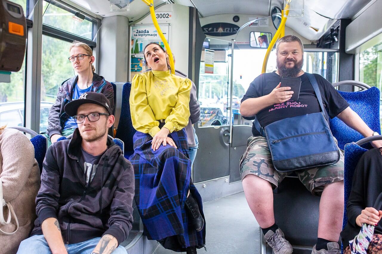 wnętrze autobusu, widać licznych pasażerów, w tym ubraną w żółtą bluzkę i niebieską spódnicę aktorkę, która siedzi z szeroko otwartymi ustami