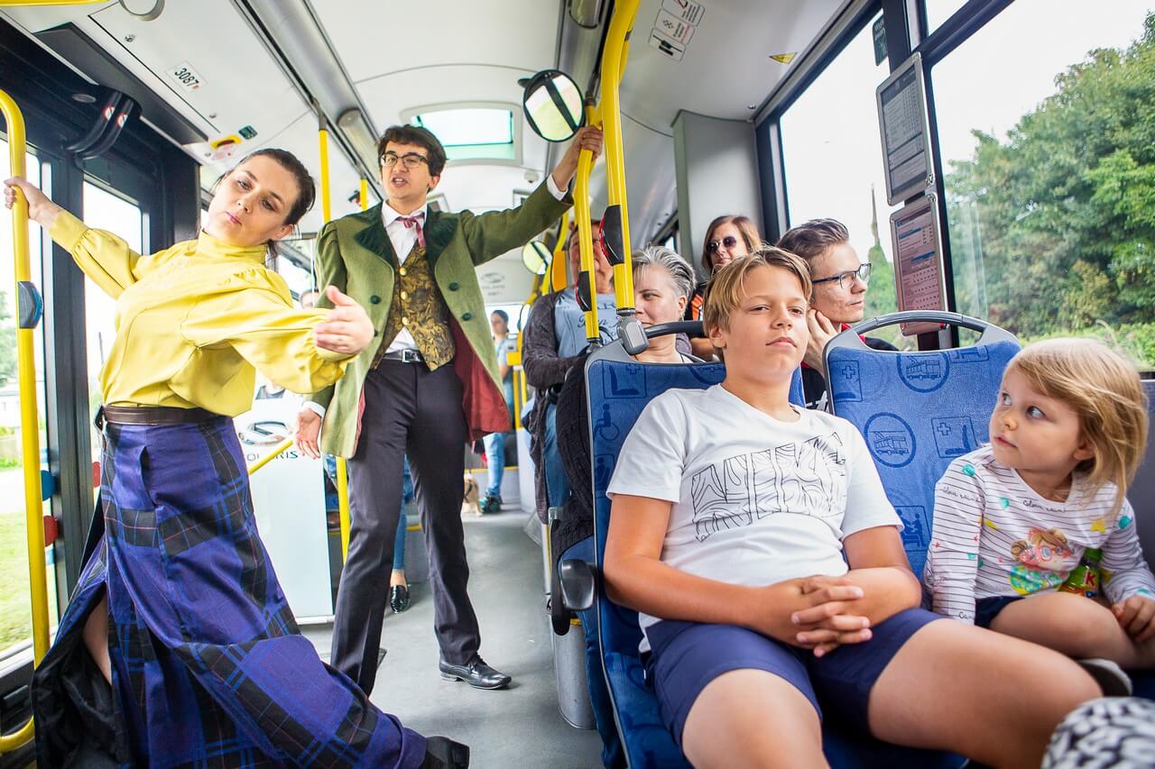 wnętrze autobusu, widać wielu siedzących pasażerów, w przejściu stoją dwie postacie, po lewej kobieta w żółtej bluzce i długiej niebieskiej spódnicy w kratę, po prawej młody mężczyzna ubrany elegancko, w zielonej marynarce i okularach, kobieta jest w trakcie dynamicznego ruchu