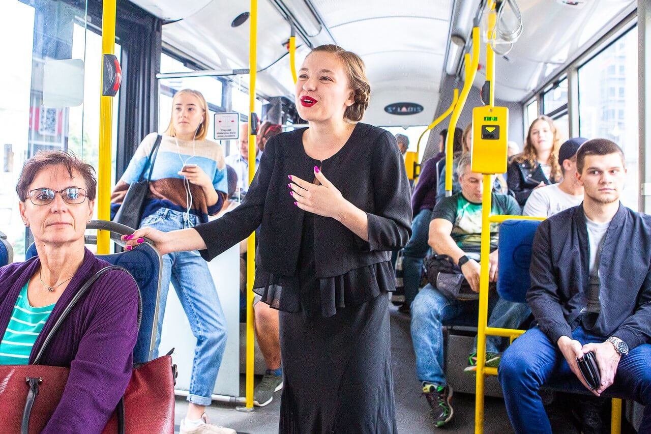 wnętrze autobusu, siedzą w nim liczni pasażerowie, na środku stoi kobieta elegancko ubrana na czarno, z czerwonymi ustami, mówi coś