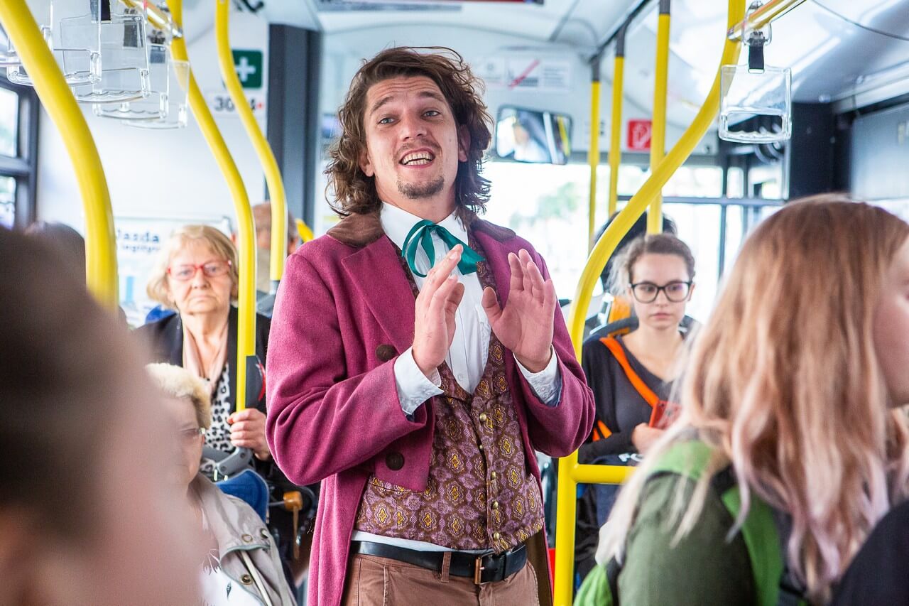 W autobusie siedzą liczni pasażerowie. Na środku stoi mężczyzna ubrany w różowy frak, kamizelkę oraz białą koszulę. Ma lekko uniesione ręce. Mówi coś.