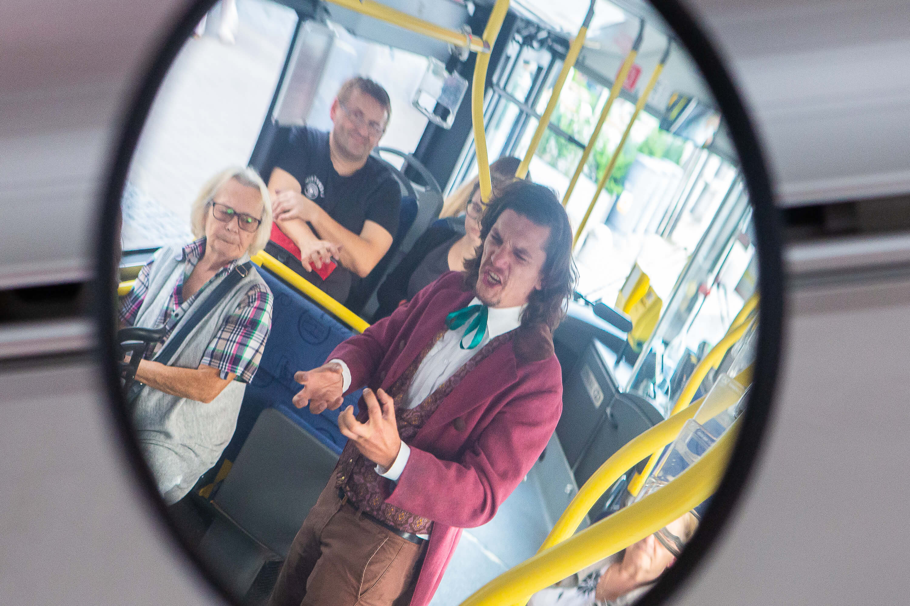 Zdjęcie odbicia w lustrze w środku autobusu. Widać na nim kilku pasażerów oraz stojącego mężczyznę ubranego w różowy frak. Intensywnie gestykuluje i mówi coś pełen pasji.