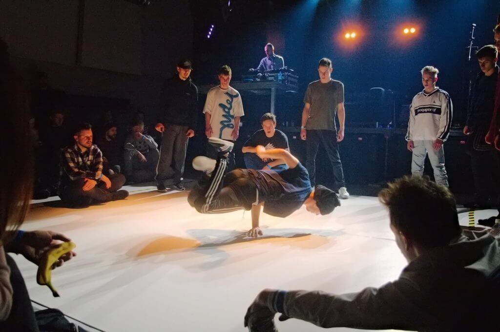 Zdjęcie z zawodów Gdynia Breaks. Na środku sceny znajduje się się tancerz, który stoi na jednej ręce. Dookoła niego stoją inni uczestnicy konkursu. Za resztą uczestników stoi DJ grający muzykę.