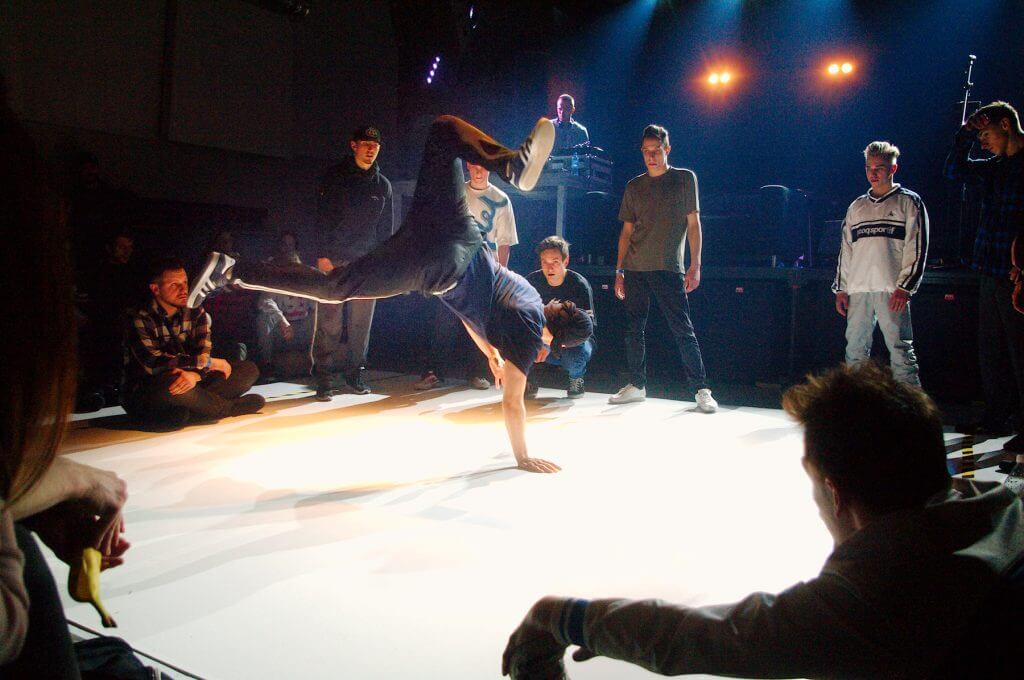 Zdjęcie z zawodów Gdynia Breaks. Na zdjęciu widać tańczącego mężczyznę, który stoi na jednej ręce i się na niej obraca. Dookoła niego są zgromadzeni inni uczestnicy konkursu.