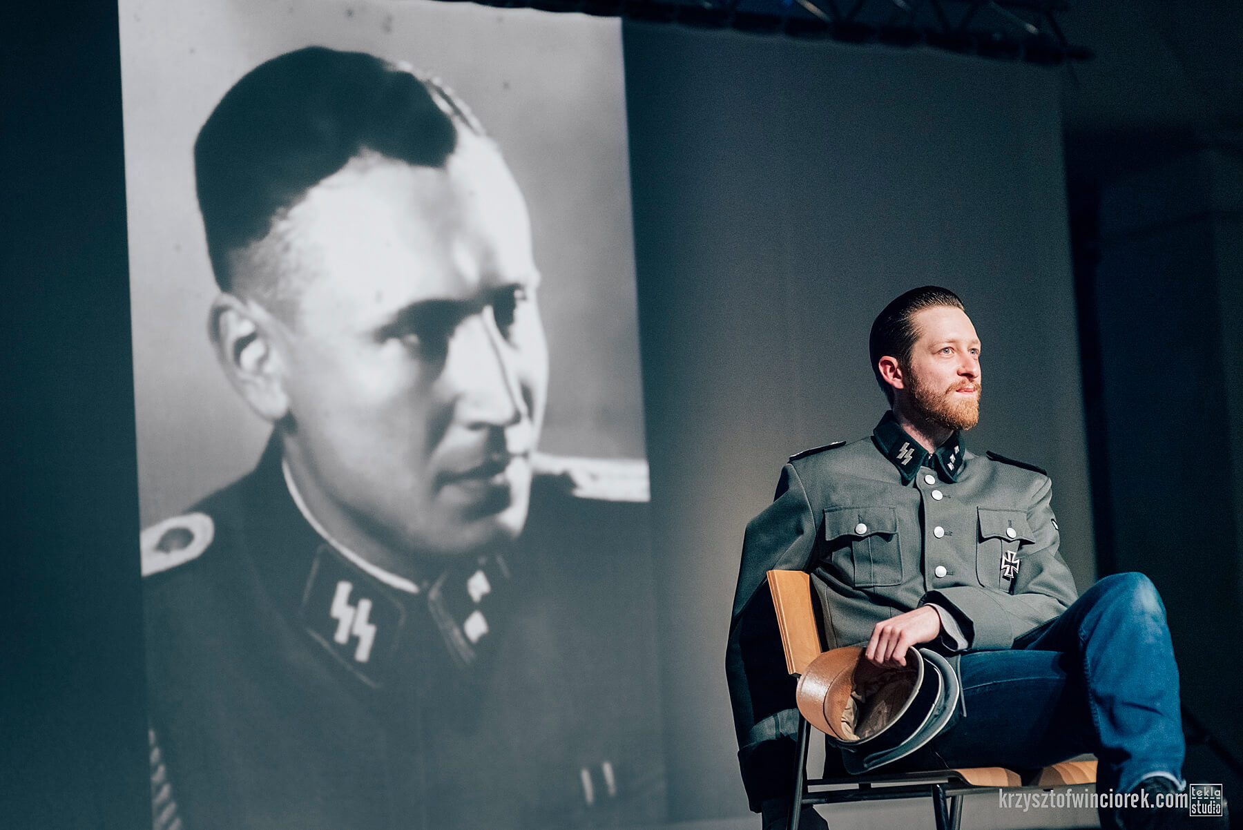 Po prawej stronie siedzi na krześle aktor z zarostem, w mundurze SS. Za nim wyświetlane jest zdjęcie historyczne jakiegoś mężczyzny, również w mundurze SS.