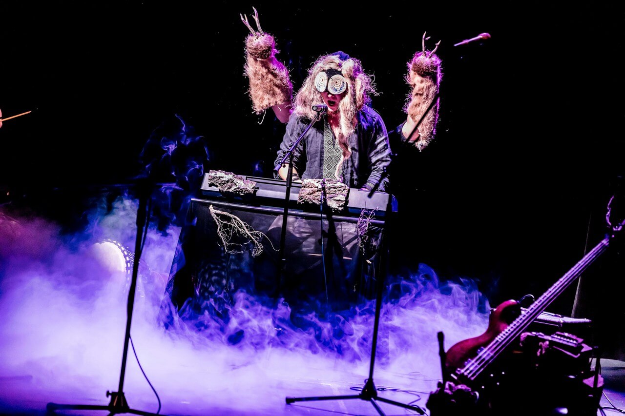 mężczyzna gra na keyboardzie, ma długie włosy, maskę na oczach, otwarte usta, przed nim ustawiony mikrofon na statywie, zza niego wystają dwie ręce w pasynkach z rogami, nad sceną unosi się dym