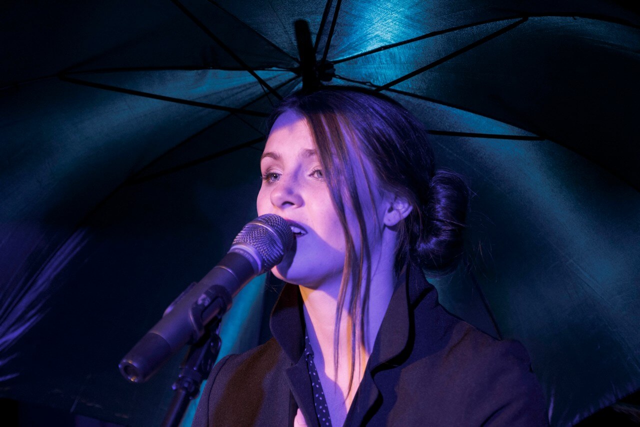 zbliżenie na popiersie kobiety, jest ubrana na czarno, włosy spięte w kok, zwrócona jest w lewo, przy ustach ma mikrofon na statywie, jest w trakcie śpiewania, stoi pod parasolem