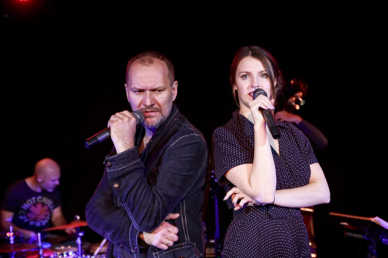 po lewej stronie mężczyzna w czarnej kurtce zwrócony w lewą stronę, w lewej ręce trzyma mikrofon blisko ust, prawą ręką podpiera łokieć, po prawej stronie kobieta w czarnej sukience, w prawej ręce trzyma mikrofon blisko ust, lewą ręką podpiera łokieć, jest w trakcie śpiewania, w te mężczyzna grający na perkusji