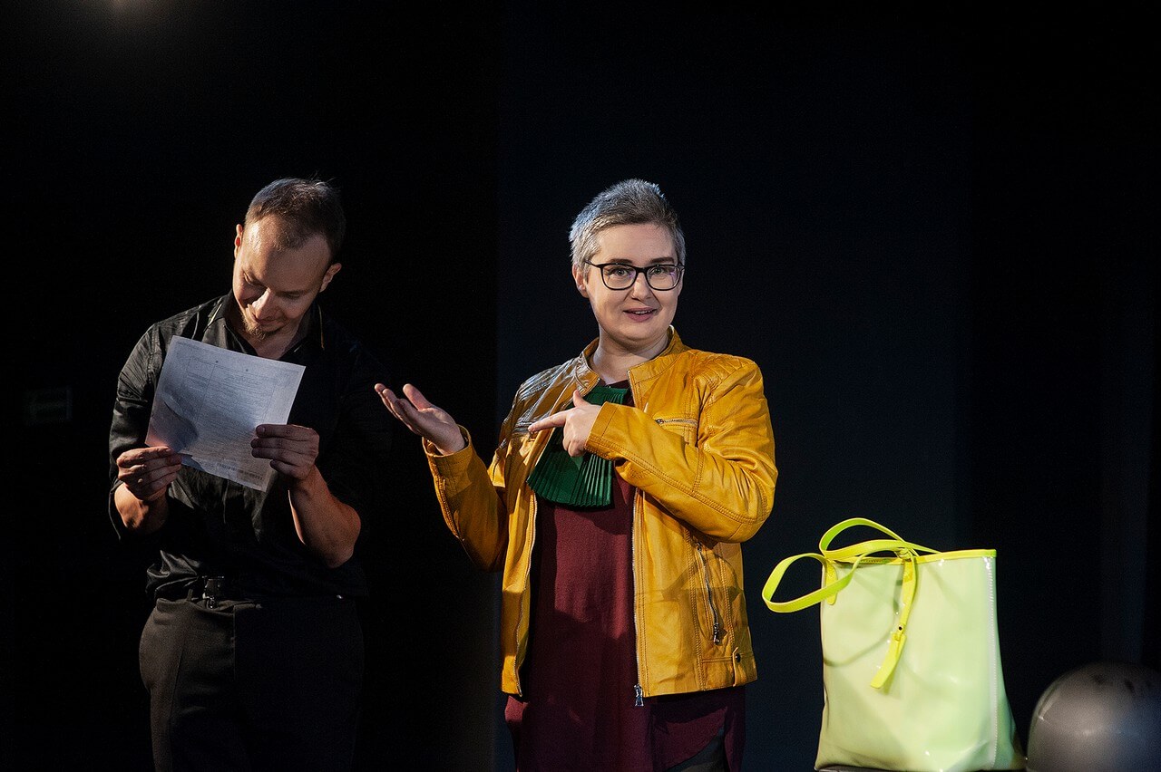dwoje aktorów stoi, mężczyzna po lewej ubrany na czarno trzyma kartkę i na nią patrzy, po prawej kobieta w okularach i żółtej kurtce patrzy przed siebie i wskazuje na mężczyznę, czarne tło