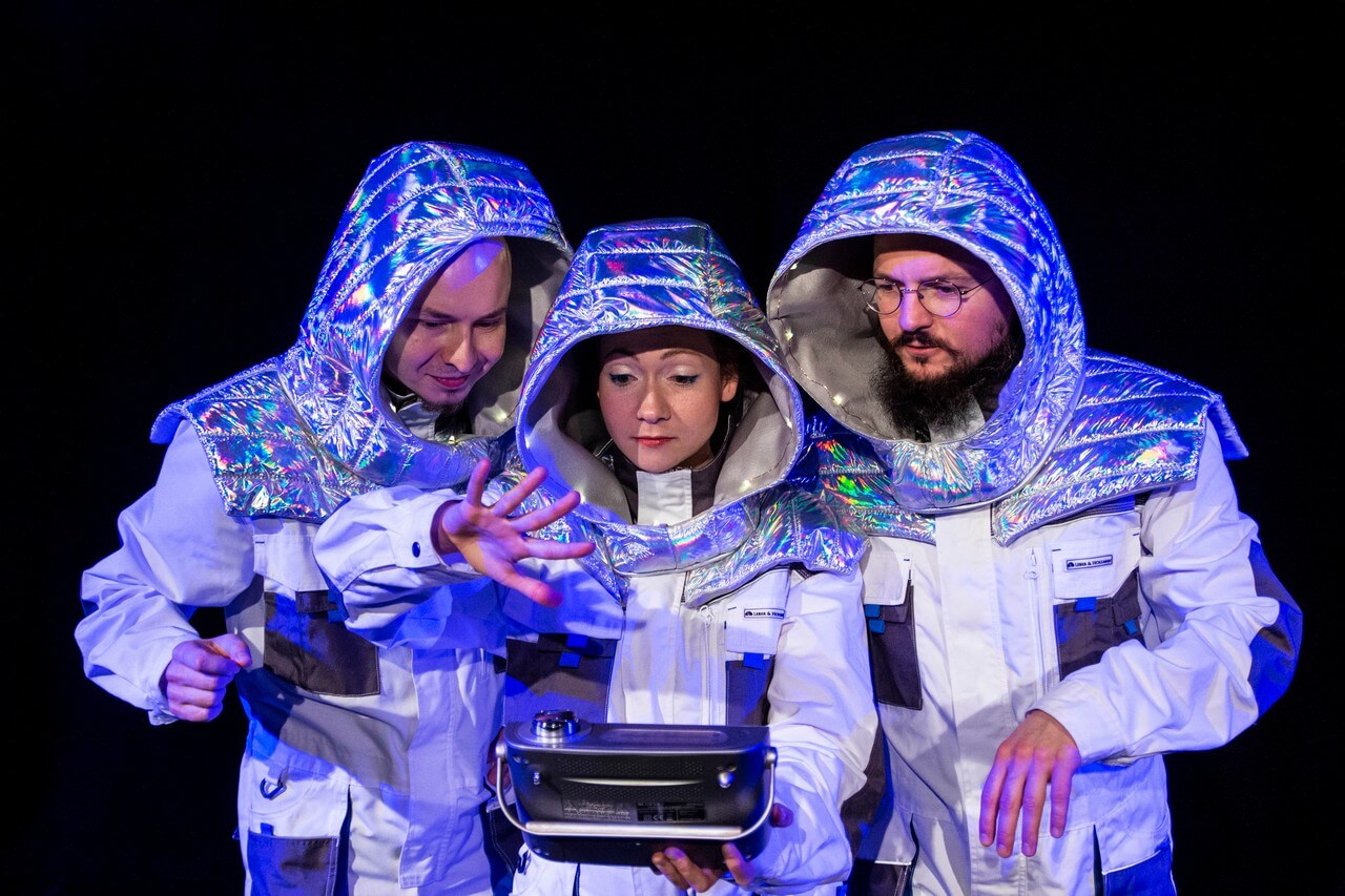 trójka aktorów w strojach astronautów, dwóch mężczyzn po bokach, kobieta w środku, patrzą na obiekt trzymany przez kobietę, kobieta unosi rękę