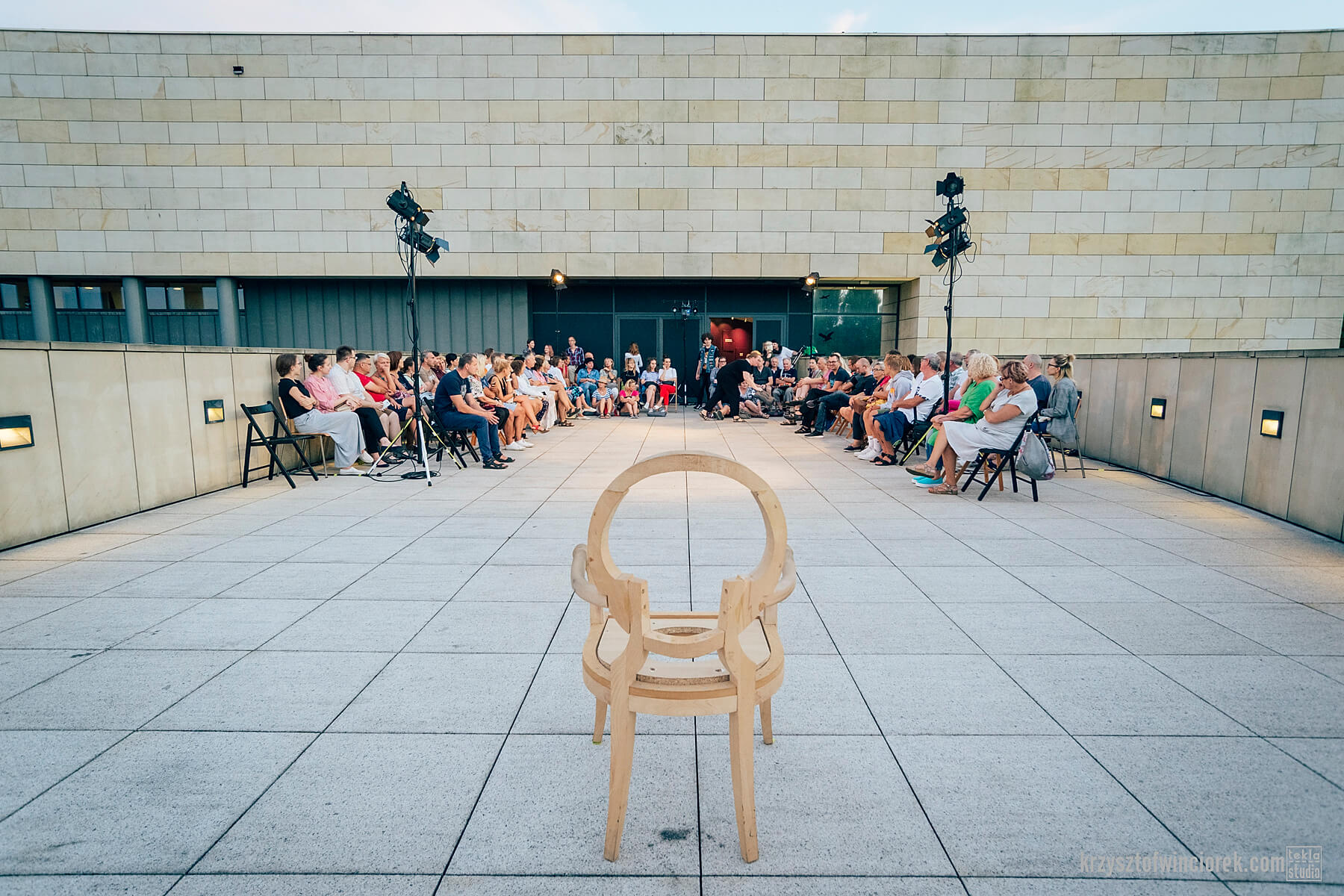 grupa widzów usadzonych w kształcie podkowy w tle, na pierwszym planie drewniane krzesło, cała scena ma miejsce na tarasie budynku Muzeum Miasta Gdyni