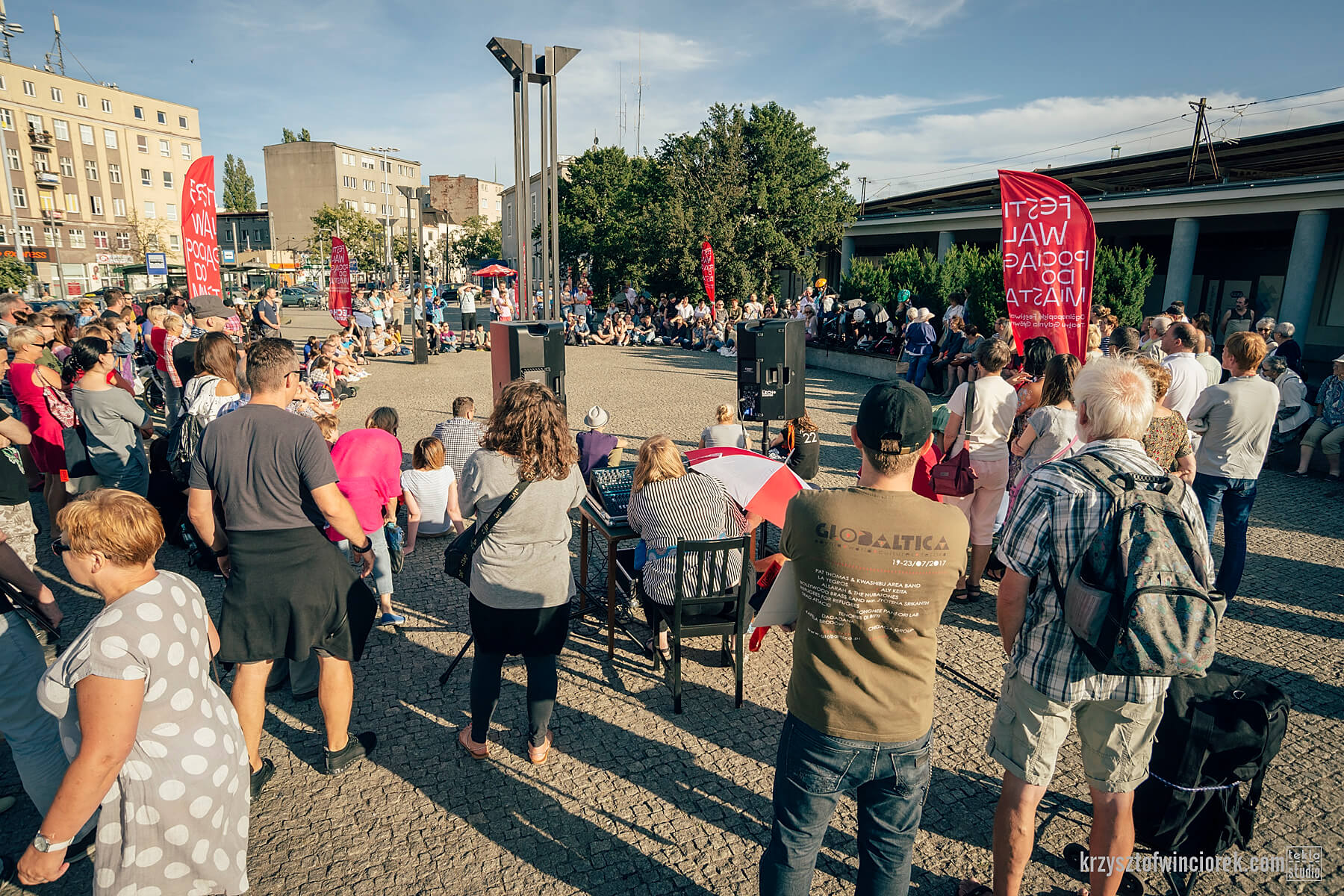 Plac przed dworcem Gdynia główna, wiele osób stoi bądź siedzi w okręgu, na zdjęciu widać ustawione nagłośnienie i banery festiwalu Pociąg do miasta