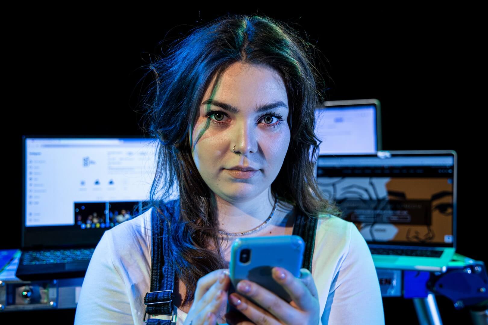 zdjęcie portretowe, kobieta w białej bluzce, z telefonem w reku, na drugim planie kilka włączonych laptopów.
