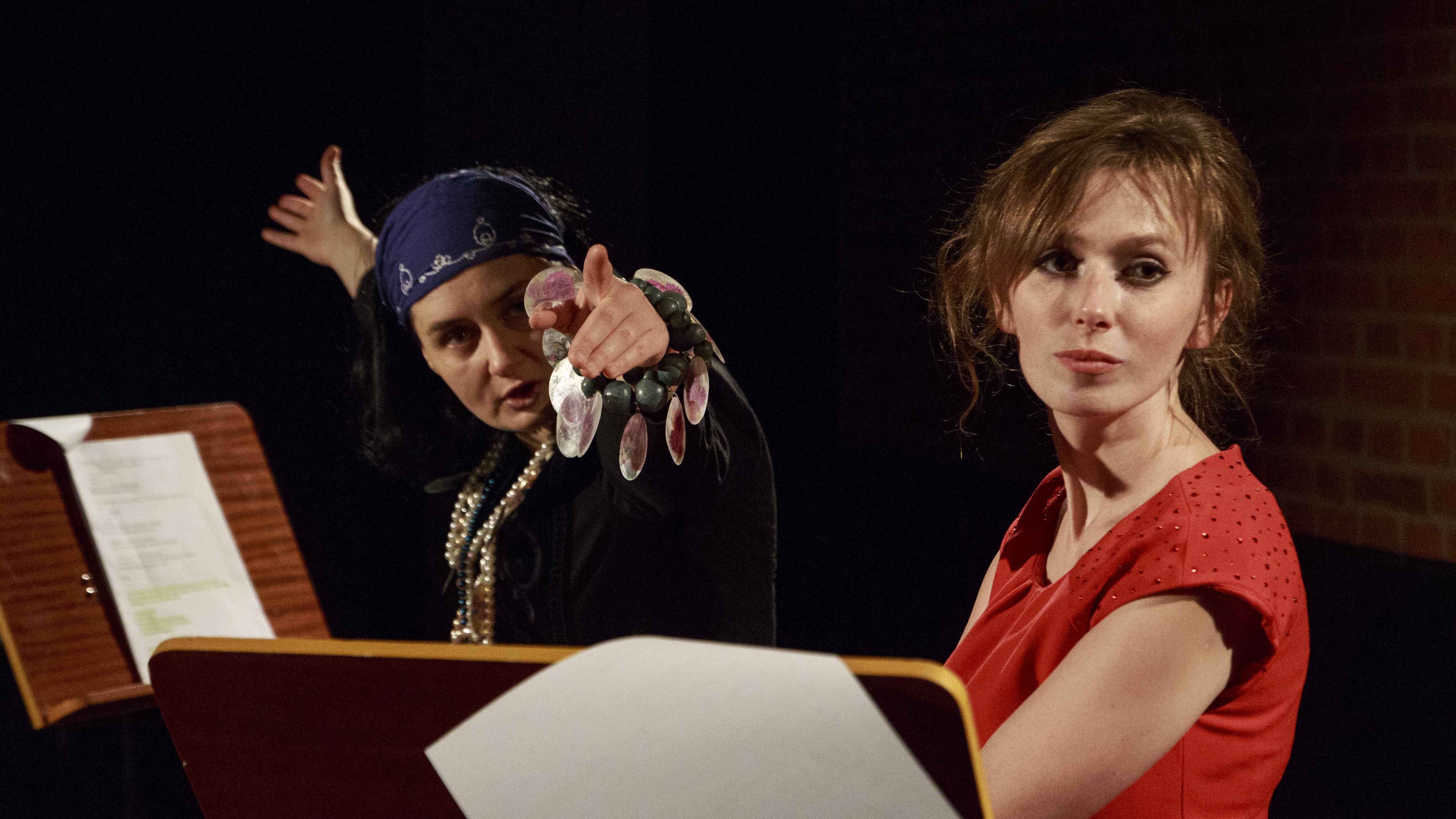 Zdjęcie ze spektaklu Dzielnice cudów. Dwie aktorki przy pulpitach z tekstem. Na pierwszym planie aktorka w czerwonej sukience, na drugim aktorka przebrana jak cyganka.