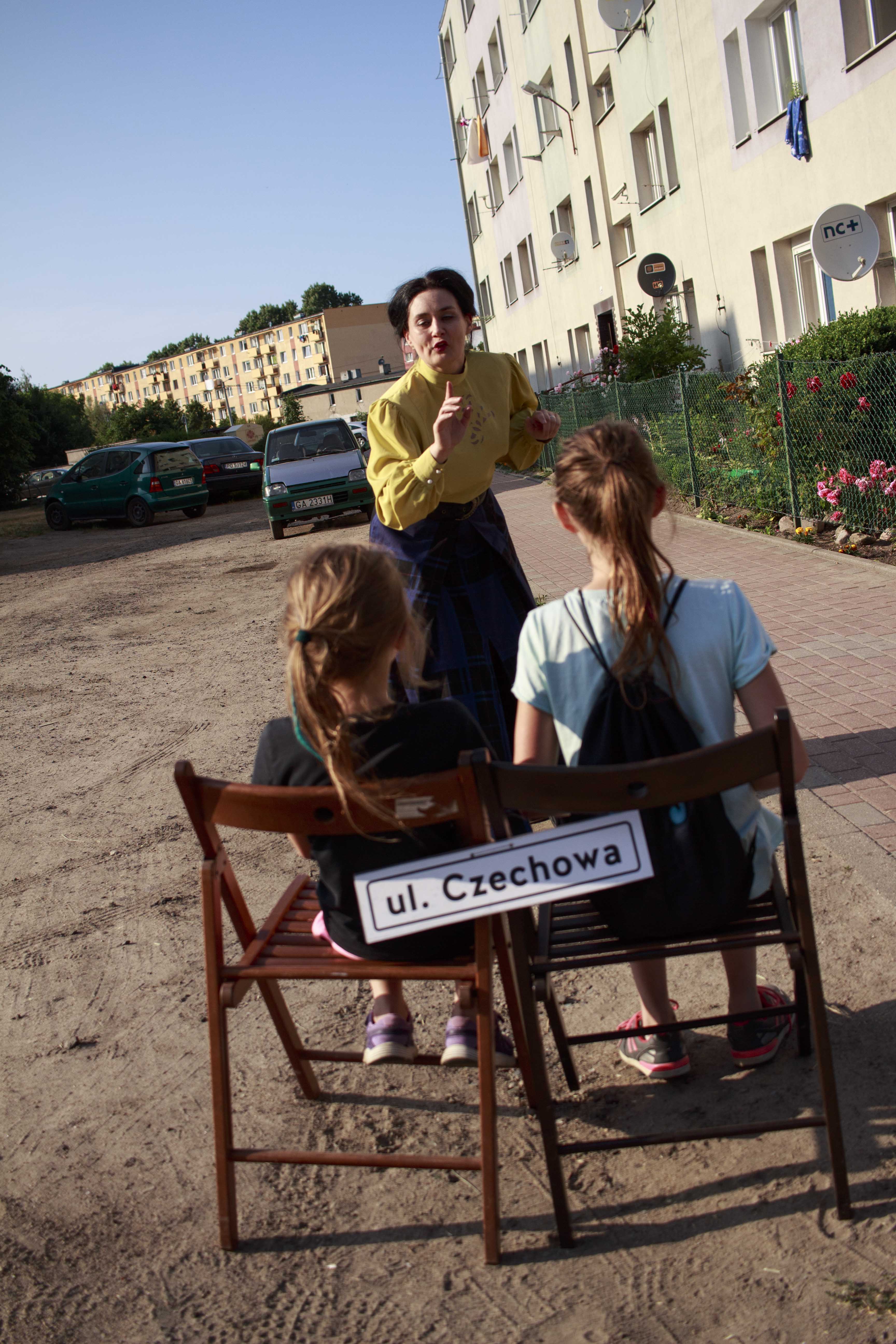 Na krzesłach z podpisem ul. Czechowa siedzą dwie dziewczynki. Przed nimi stoi kobieta ubrana w długą, ciemną spódnicę i żółtą koszulę. Kobieta pochyla się w kierunku dziewczynek.