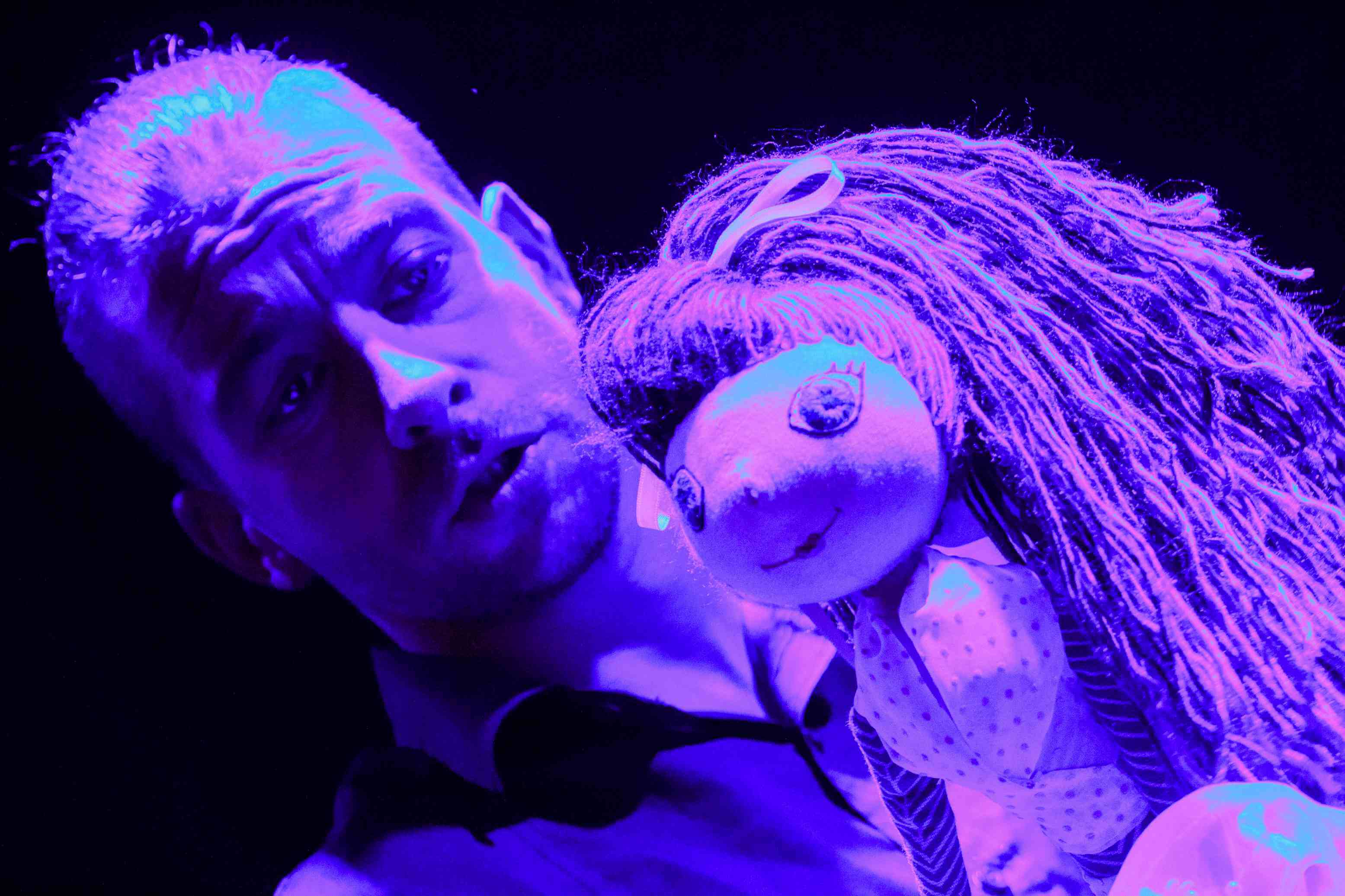 mężczyzna animuje lalkę, lalka ma jasną sukienkę i niebieskie włosy, zbliżenie na lalkę, widać ją od pasa w górę, czarne tło, pada intensywne fioletowe światło