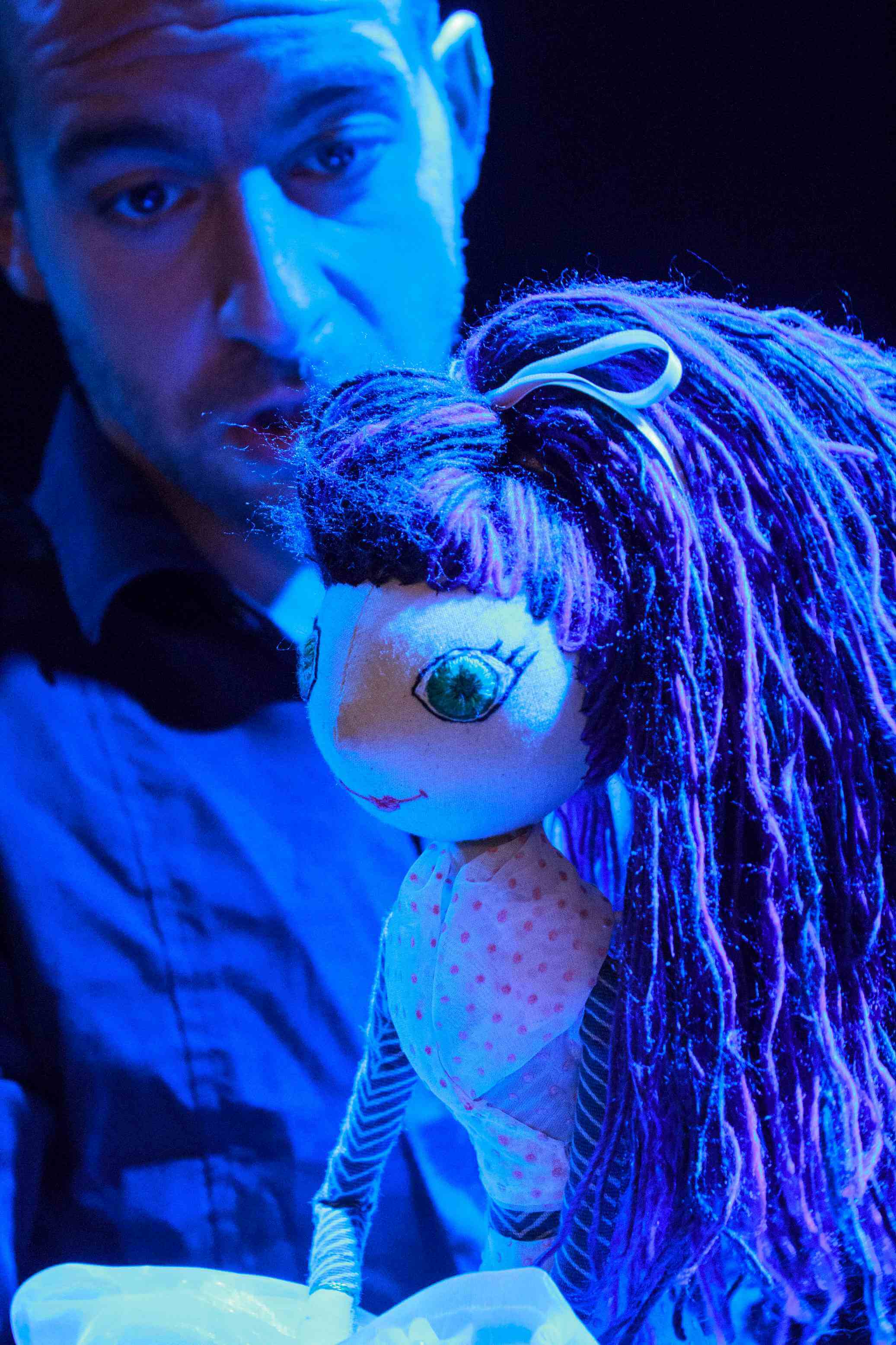 ubrany na czarno mężczyzna animuje lalkę w jasnej sukience i z niebieskimi włosami, zbliżenie na lalkę, lalka ma zielone oczy, czarne tło, pada niebieskie światło