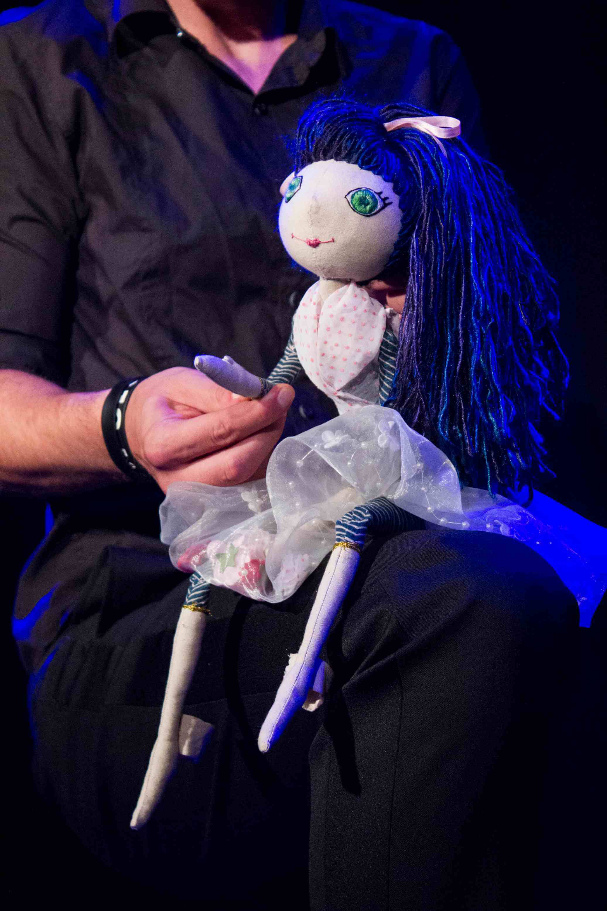 lalka siedząca na udzie animującego ją mężczyzny ubranego na czarno, mężczyzna trzyma w dłoni jedną z rąk lalki, lalka ma jasną sukienkę i niebieskie włosy, zielone oczy, czarne tło, pada niebieskie światło