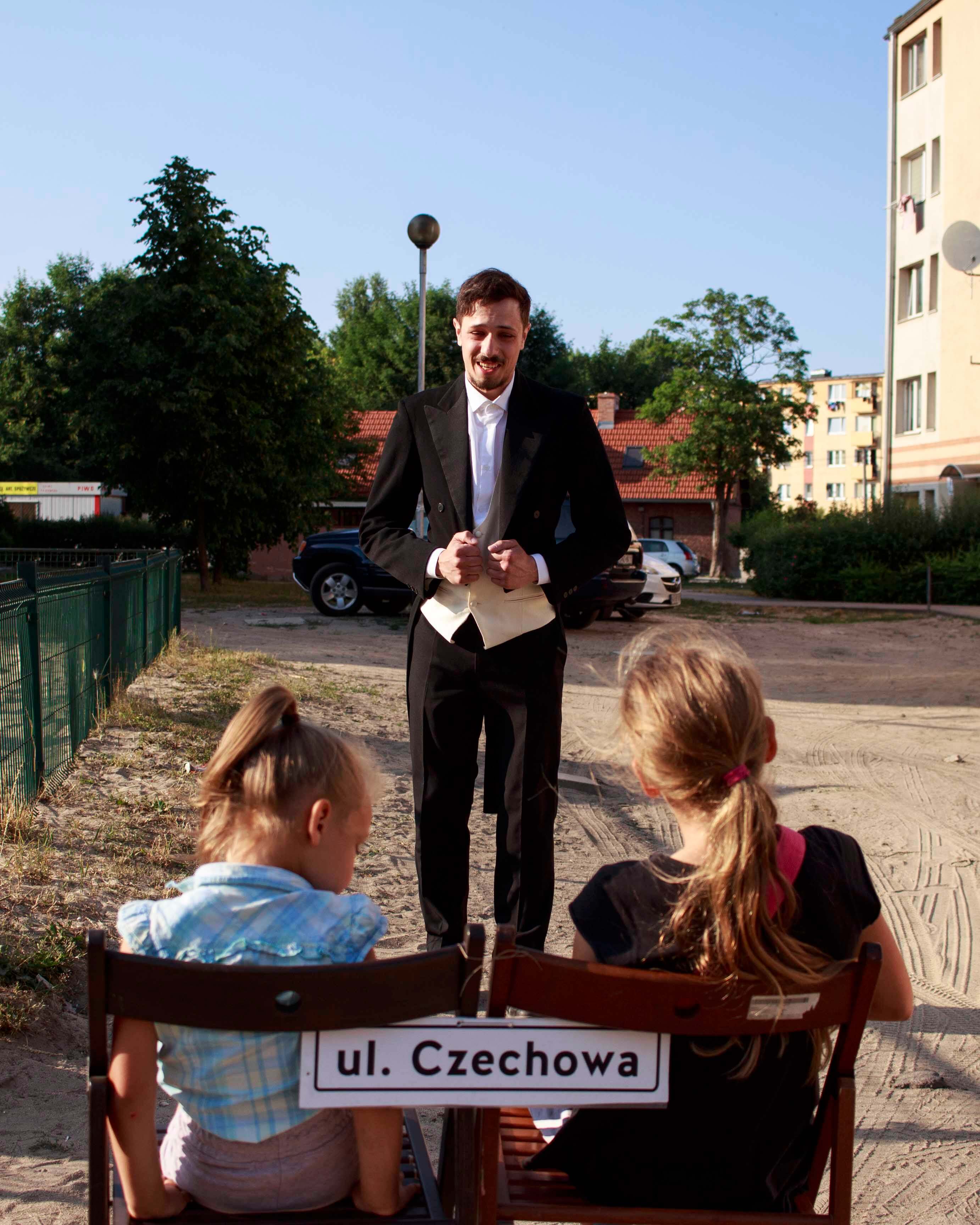 Dwie dziewczynki siedzą na krzesłach z podpisem ul. Czechowa. Przed nimi stoi uśmiechnięty mężczyzna we fraku.