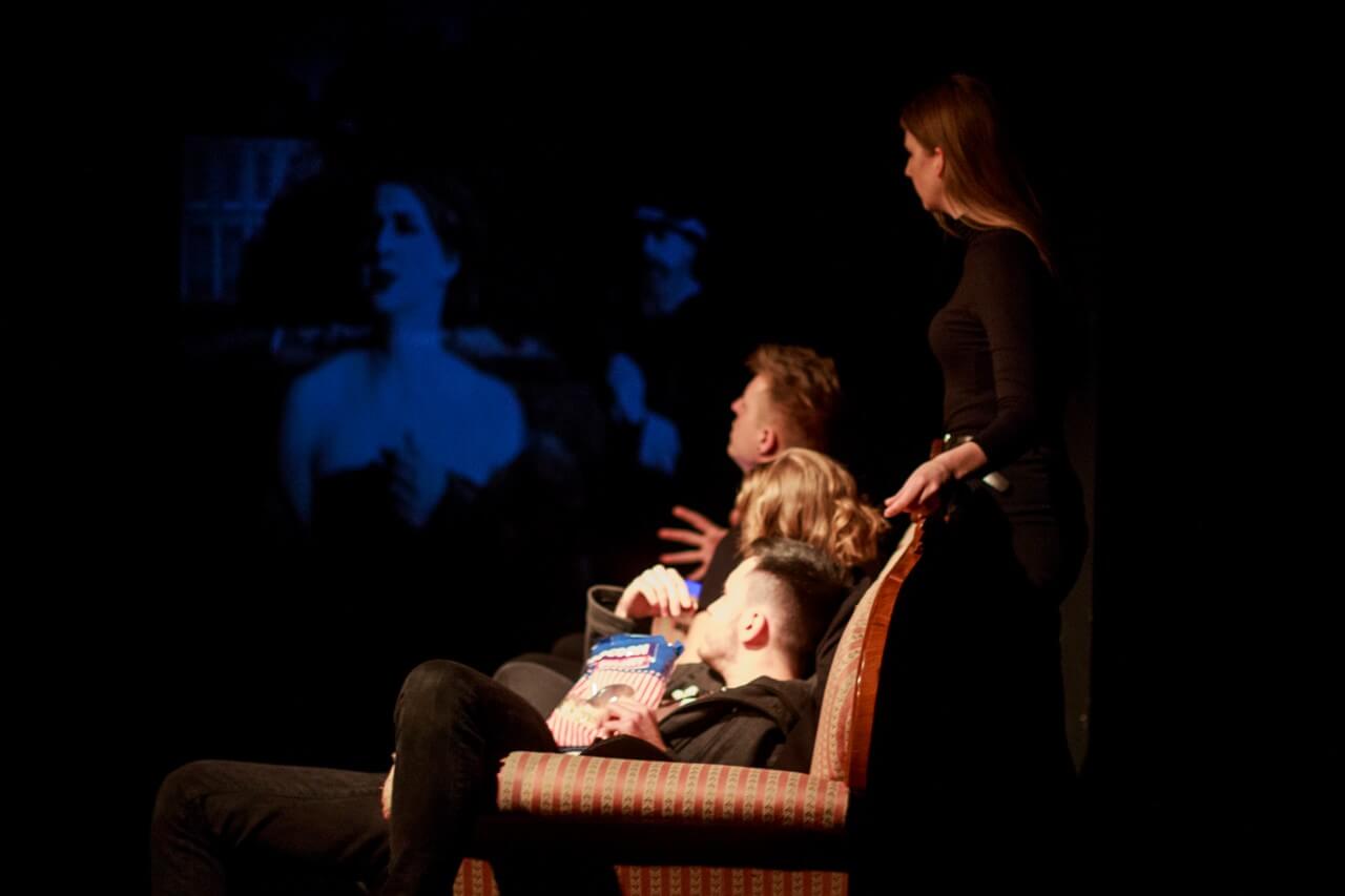 czworo aktorów ubranych na czarno, dwie kobiety i dwóch mężczyzn, troje siedzi na kanapie w paski i je popkorn, za kanapą stoi kobieta i opiera dłonie o oparcie, wszyscy patrzą w bok na wyświetlany obraz przedstawiający kobietę
