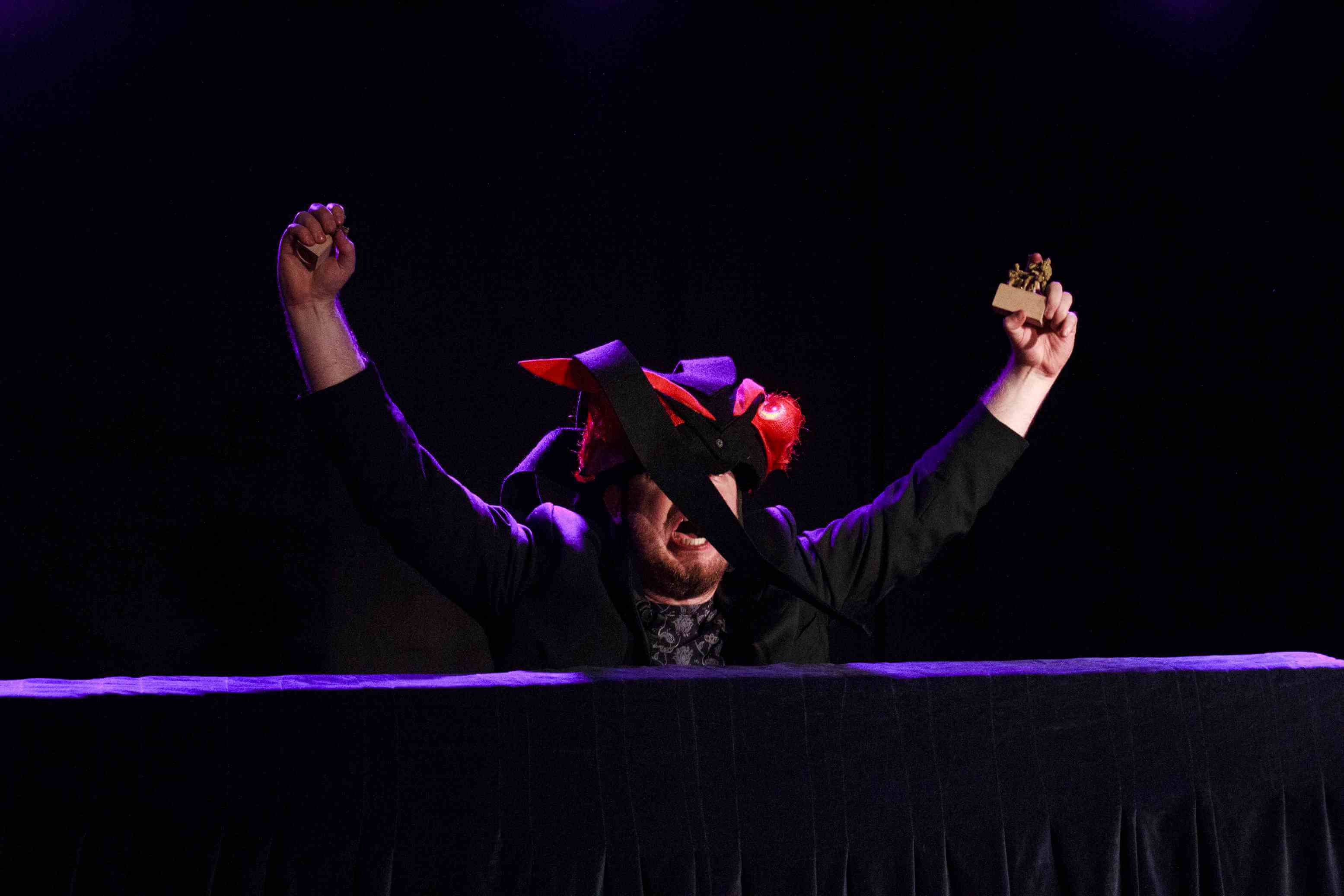 aktor wyłania się zza czarnego blatu, ubrany jest na czarno, na głowie ma makietę głowy czarengo smoka z czerwonymi oczami, aktor ma otwarte usta, krzyczy, ręce ma uniesione do góry, w dłoniach trzyma zabawkowe żołnierzyki ciemne tło