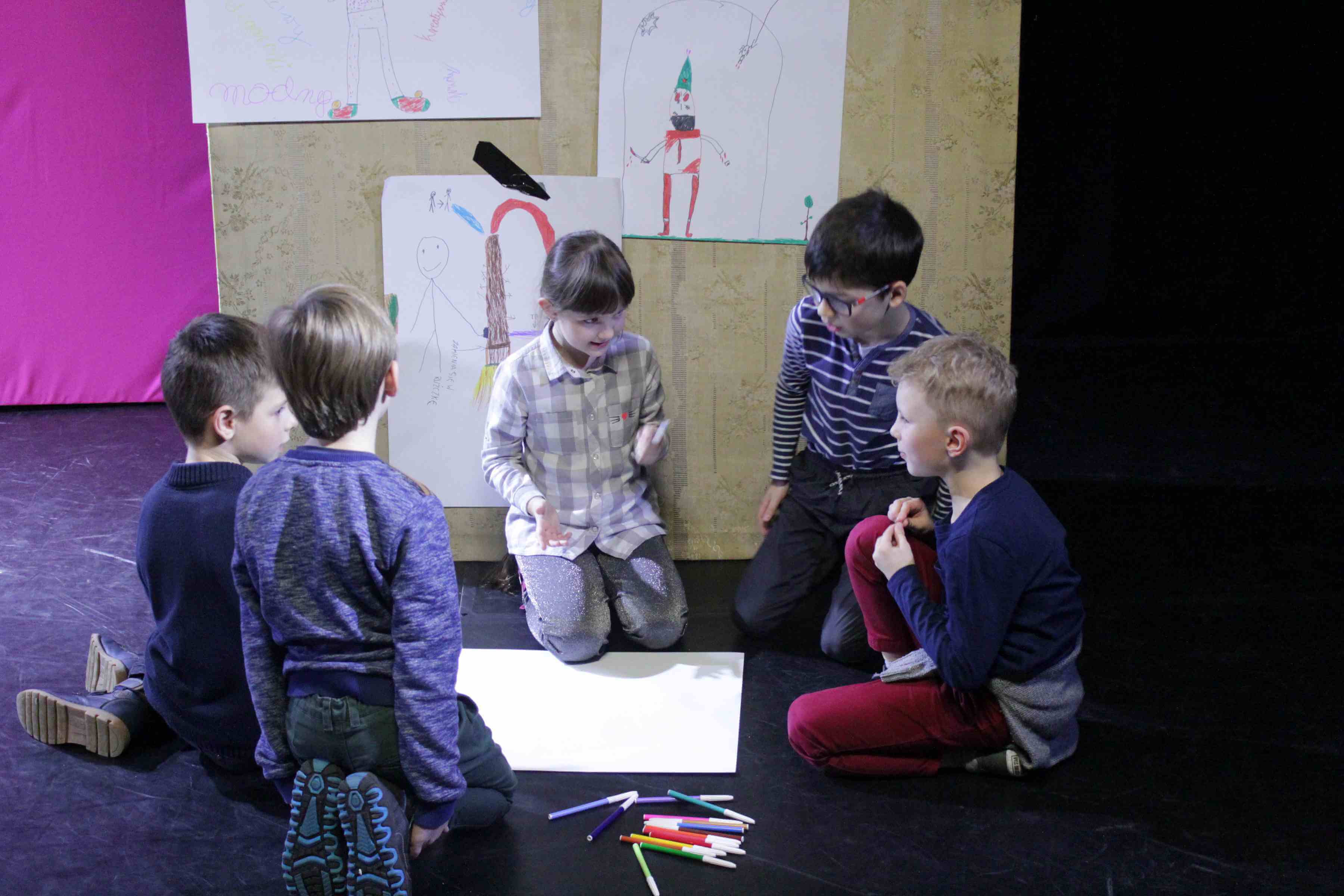 krupka dzieci siedzi na podłodze, obok nich leżą przygotowane kolorowe flamastry, między nimi na podłodze leży kartka papieru. w tle widaćpłytę drewnianą z pracami plastycznymi innych dzieci, czarna podłoga, po lewej stronie różowe tło, po prawej czarne