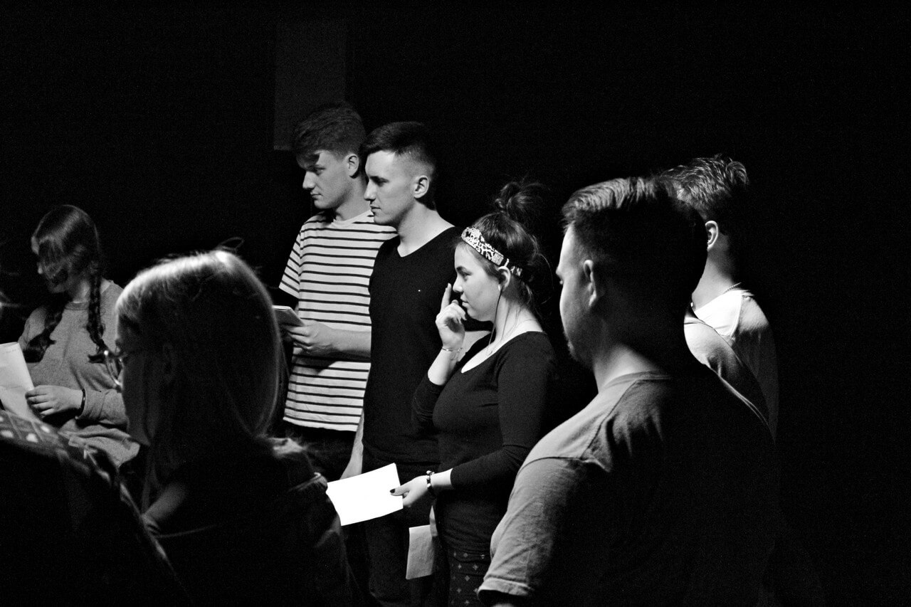 czarnobiałe zdjęcie, grupa osób w młodym wieku stoi obok siebie, trzymają w dłoniach kartki, niektórzy na nie spoglądają, inni dwoma palcami wolnej ręki trzymają nos, zwróceni są w lewo, tło jest ciemne, zdjęcie z wydarzenia teatru gdynia główna TEATR STUDENCKI