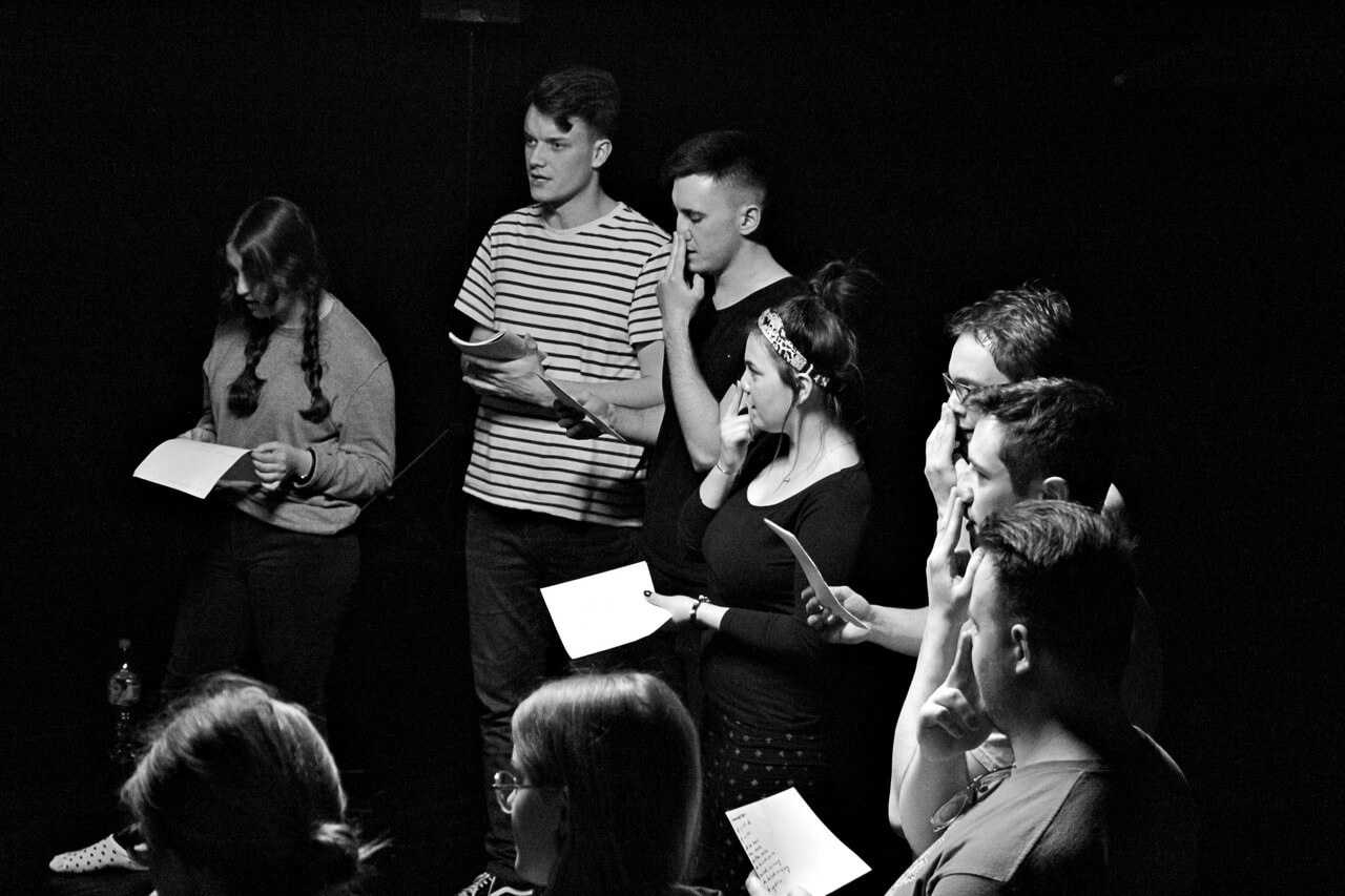 czarnobiałe zdjęcie, grupa osób w młodym wieku stoi obok siebie, trzymają w dłoniach kartki, niektórzy na nie spoglądają, inni dwoma palcami wolnej ręki trzymają nos, zwróceni są w lewo, tło jest ciemne, zdjęcie z wydarzenia teatru gdynia główna TEATR STUDENCKI