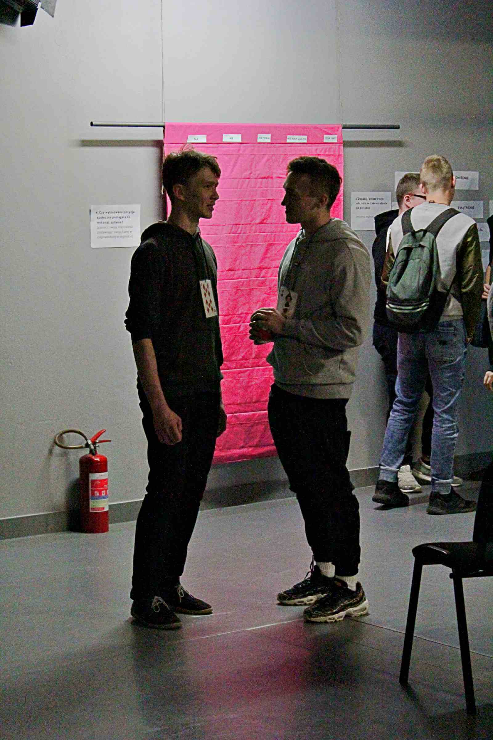 dwóch młodych mężczyzn stoi i rozmawia ze sobą, za nimi widoczne dwie osoby, na ścianie wisi czerwony materiał z kieszonkami