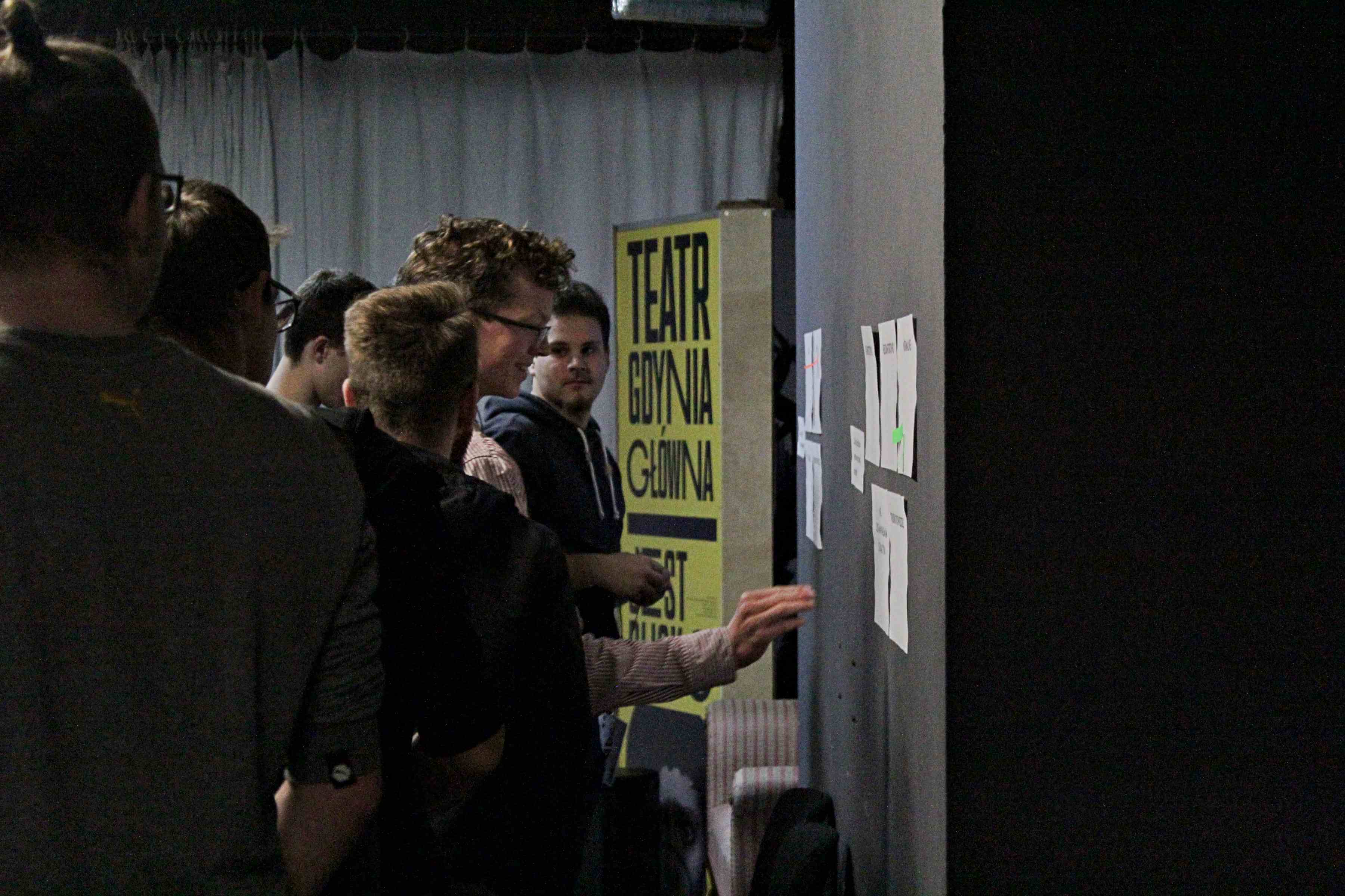 grupa młodych ludzi stojąca przy ścianie, jeden z nich przykleja coś do kartki wiszącej na ścianie, w tle napis teatr gdynia główna