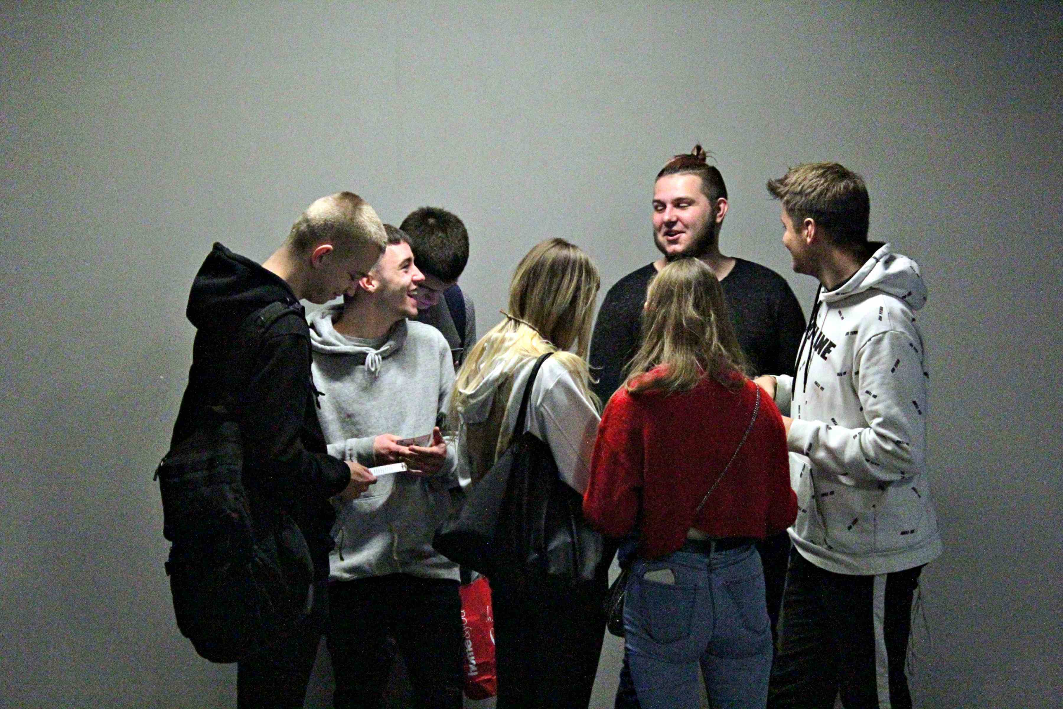 grupa młodych ludzi stoi na środku kadru na tle szarej ściany, po prawej chłopak ubrany na czarno w bluzie z kapturem i czarnym plecakiem na prawym ramieniu, obok niego chłopak w szarej bluzie z kaprurem i czarnych spodniach, za nim chłopak w szarej bluzie z czarnym plecakiem, na środku dziewczyna z długimi blond włosami w białej bluzie z kapturem, z czarną torebką na prawym ramieniu, ma czarne spodnie, obok niej dziewczyna w czerwonej bluzie i jasnych jeansach, za nią chłopak w czarnek koszulce, obok niego chłopak w biało czarnej bluzie z kapturem i czarnych spodniach z białym lampasem,
