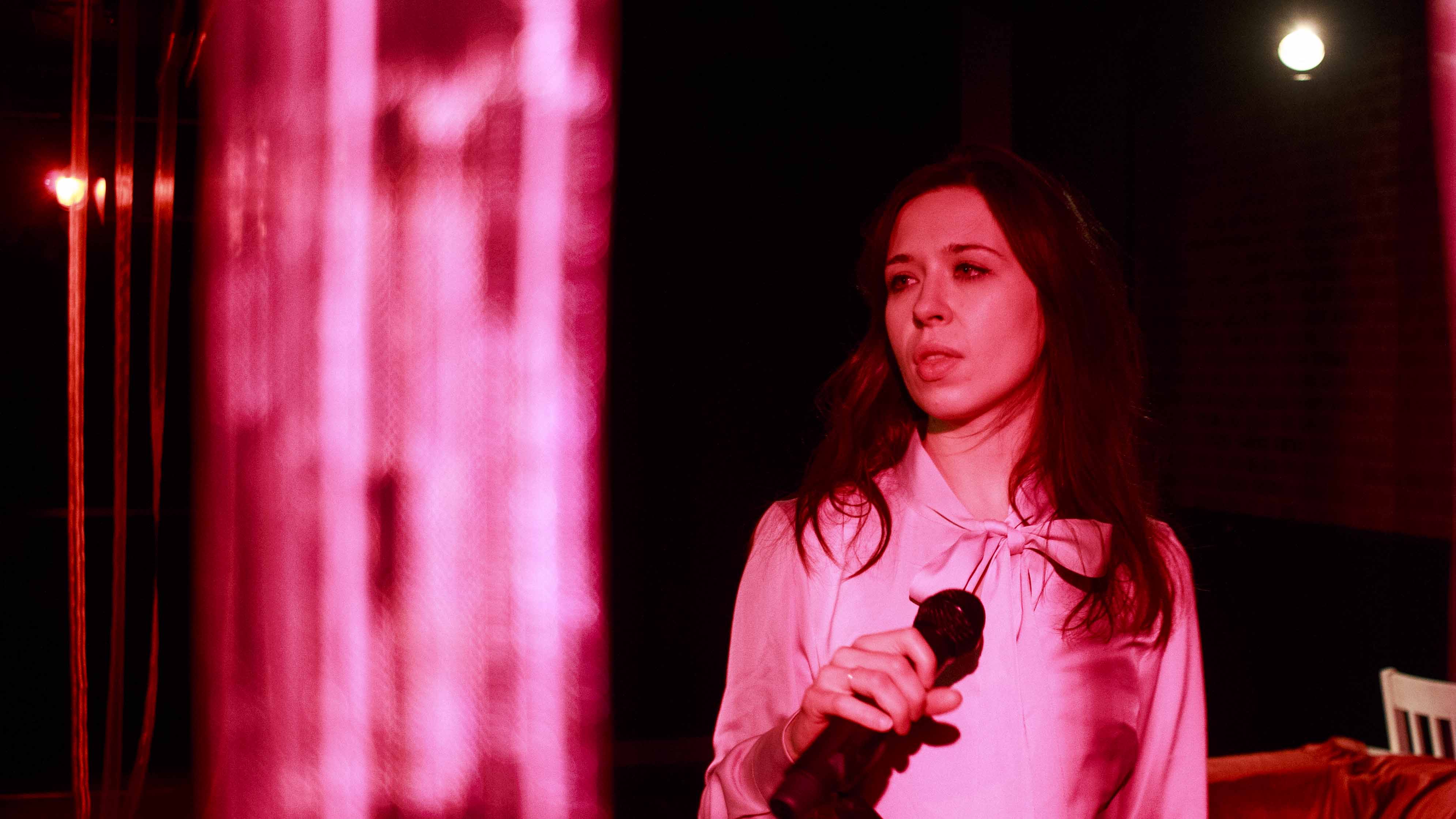 Zdjęcie do spektaklu Dobroć nasza dobroć. Aktorka w rózowo czerwonym świetle trzyma mikrofon.