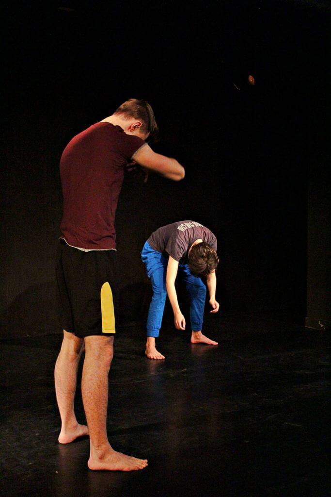 Zdjęcie z projektu Teatr Młodych. Uczestnicy ćwiczą na czarnej sali teatralnej. Jeden kuca ze zwieszoną w dół głową, a drugi chowa głowę między swoje ręce.