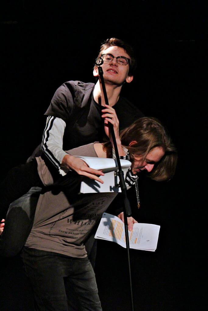 Zdjęcie z projektu Teatr Młodych. Dwóch uczestników. Dwaj młodzi mężczyźni. Jeden z mężczyzn stoi na podłodze i patrzy w scenariusz, który trzyma w swojej lewej ręce, podczas gdy drugi mężczynza siedzi na jego plecach i wygłasza kwestię na wysoko postawionym mikrofonie.