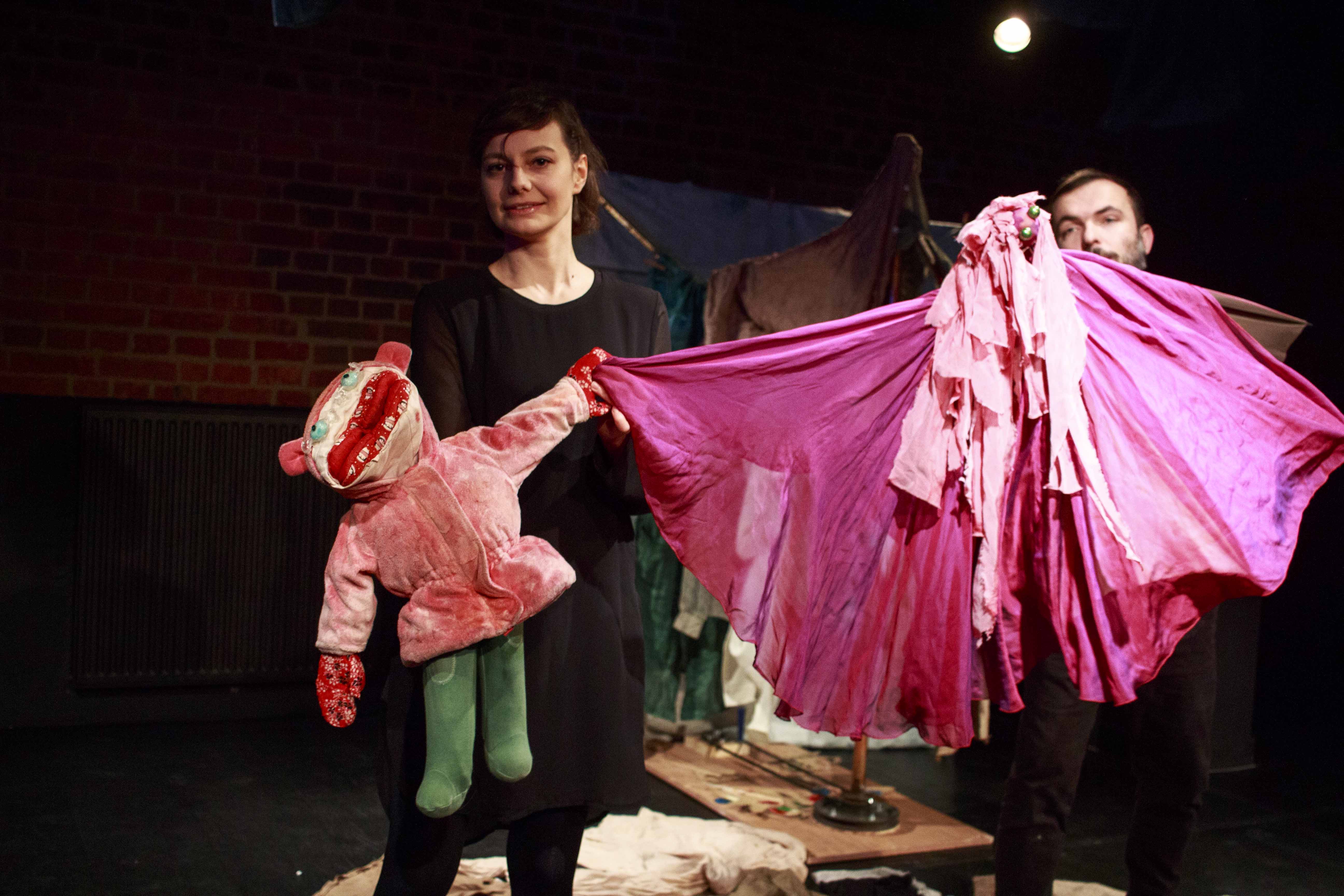 Zdjęcie do spektaklu Czerwony Kapturek (trochę inna historia). Na scenie dwóch aktorów. Aktorka trzyma w rękach kukiełkę czerwonego kapturka, któa trzyma swoją rączką sukienkę innej kukiełki, ubraną w różową sukienkę, którą trzyma drugi aktor.