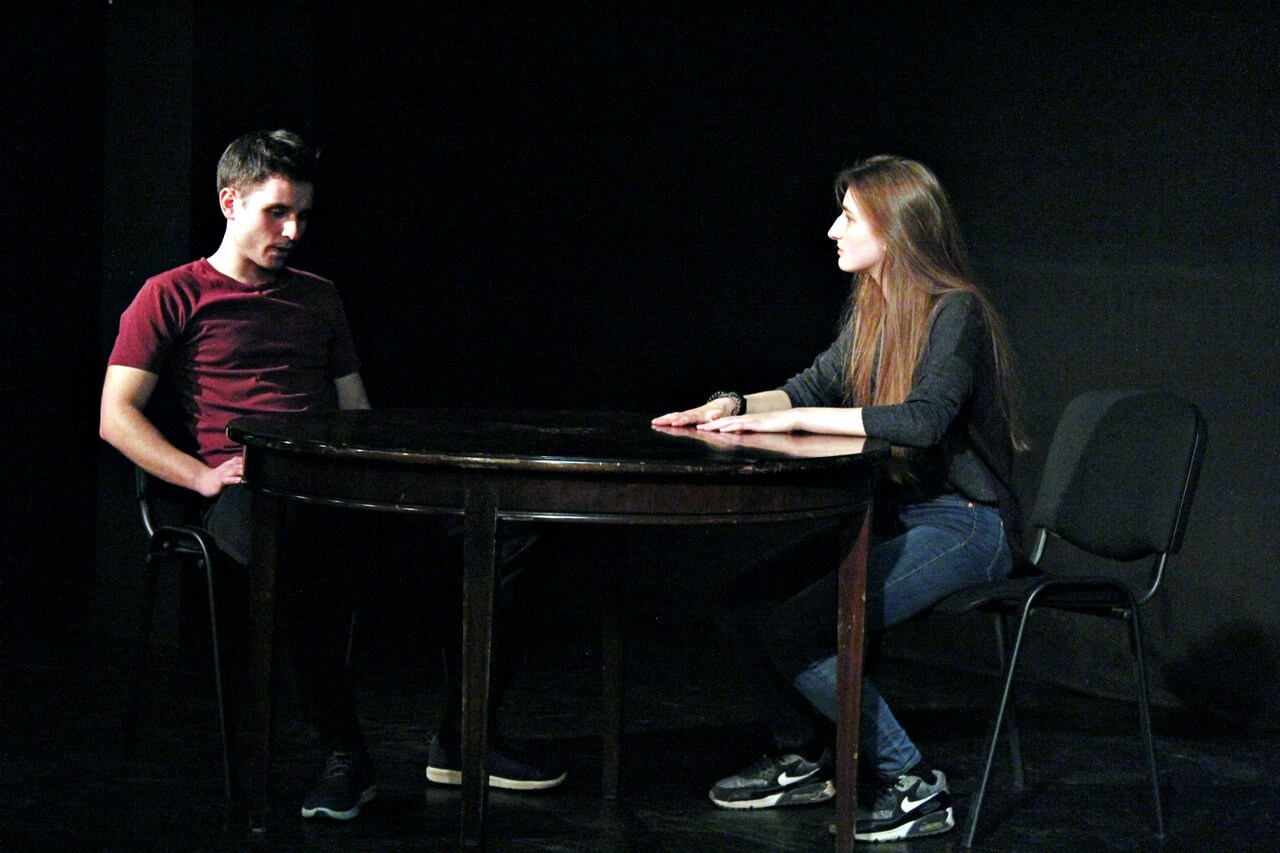 na scenie ustawiony ciemny owalny stół i dwa czarne krzesła, po lewej siedzi mężczyzna o krótkich włosach w czerwonej bluzce, ręce trzyma na kolanach, patrzy w dół, na przeciwko siedzi kobieta w długich rozpuszczonych włosach, patrzy na mężczyznę, dłonie trzyma na stole, tło ciemne, zdjęcie z wydarzenia teatru gdynia główna TEATR STUDENCKI