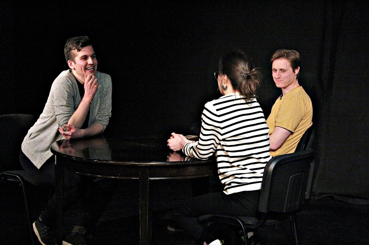 na scenie trzy osoby, na środku ustawiony ciemny owalny stolik i trzy ciemne krzesła, po lewej stronie siedzi mężczyzna w zielonej bluzce, szarym swetrze i ciemnych spodniach, uśmiecha się, jedno ramie opiera o blat stołu drugą dłoń trzyma na brodzie, obok siedzi mężczyzna w jasnych krótkich włosach i żółtej bluzce, patrzy na siedzącą obok kobietę we włosach spiętych w kucyk i bluzce w biało czarne paski, opiera ona dłonie na stole, ciemne tło, zdjęcie z wydarzenia teatru gdynia główna TEATR STUDENCKI