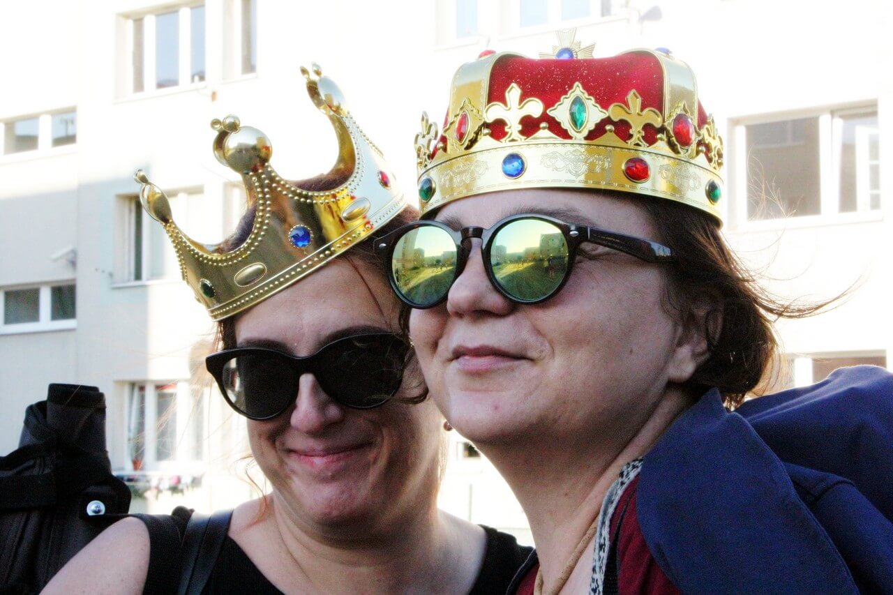 przestrzeń osiedla mieszkalnego, zbliżenie na twarze dwóch kobiet, obie mają okulary przeciwsłoneczne na oczach i złote korony na głowach