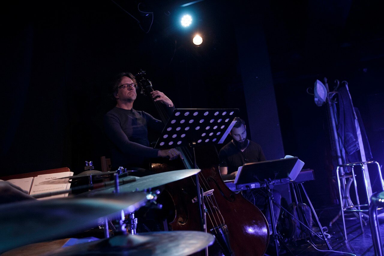 po lewej stronie mężczyzna grający na kontrabasie, po prawej mężczyzna grający na keyboardzie, po lewej stronie widoczny fragment perkusji, ciemne tło, pada niebieskie światło