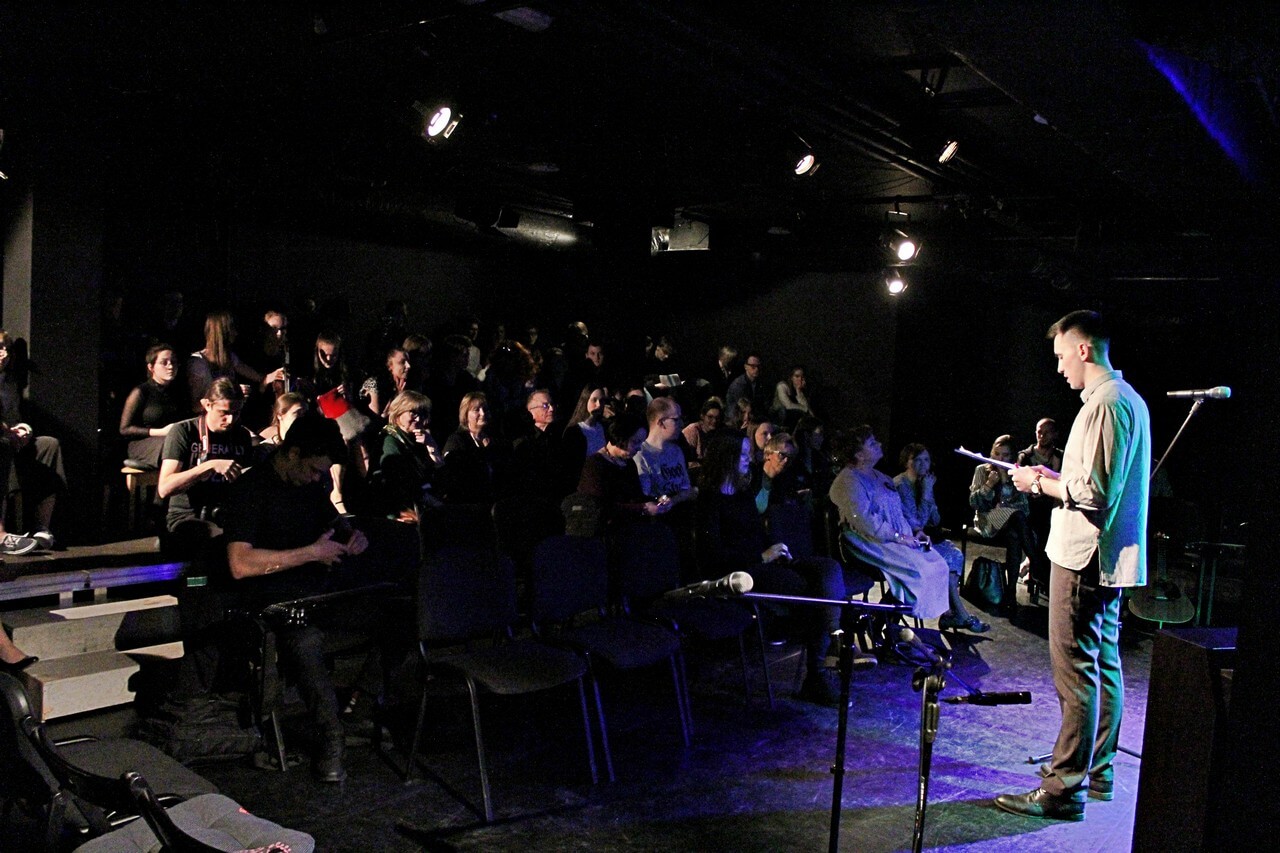 na scenie stoi mężczyzna ubrany w jasną koszulę, ciemne spodnie i eleganckie buty, trzyma w dłoniach kartkę na którą patrzy, przed nim znajdują się widzowie na widowni, na scenę pada niebieskie światło