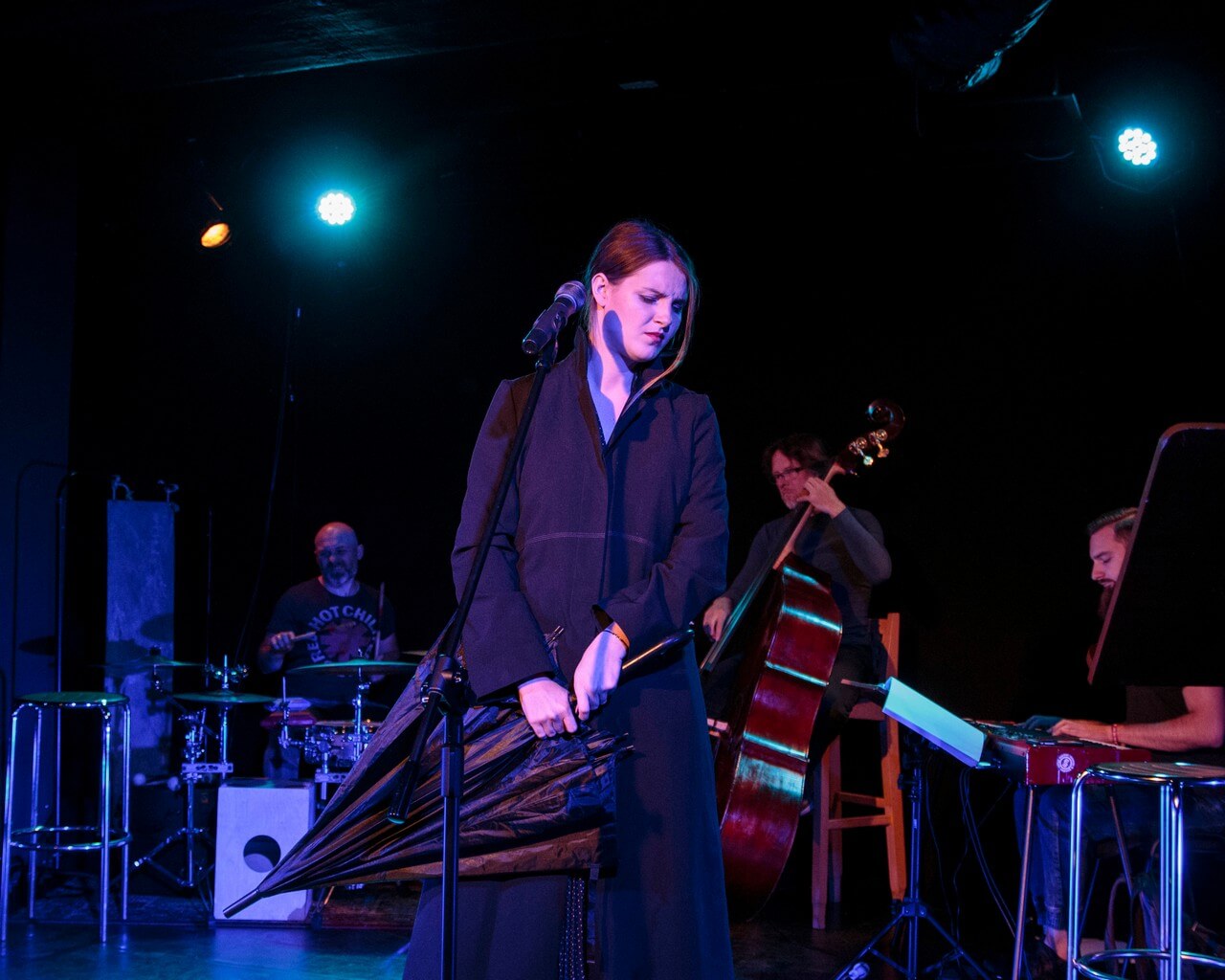 kobieta ubrana w czarną sukienkę stoi na środku, ma zamknięte oczy, zwrócona jest w prawo, w dłoniach trzyma złożony czarny parasol, przed nią stoi mikrofon na statywie, w tle po lewej mężczyzna grający na perkusji, obok mężczyzna grający na kontrabasie, po prawej stronie mężczyzna grający na keyboardzie, ciemne tło, pada niebieskie światło
