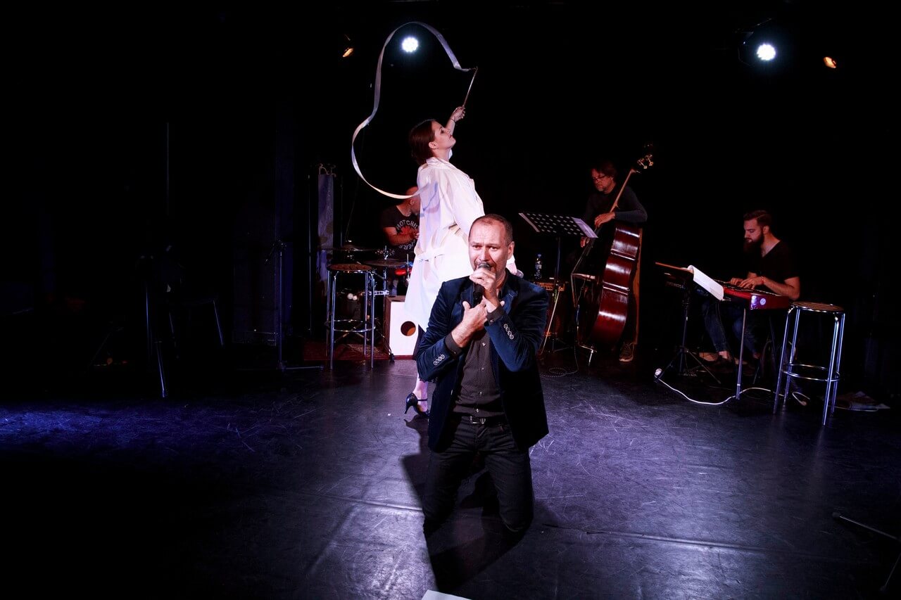 mężczyzna w niebieskiej marynarce klęczy na środku sceny, w lewej ręce trzyma mikrofon blisko ust, prawą rękę trzyma przy klatce piersiowej, za nim stoi kobieta w białej sukience, zwrócona jest w prawo, lewą rękę ma uniesioną do góry i trzyma w niej białą wstążkę, w tle widać mężczyznę grającego na perkusji, mężczyznę grającego na kontrabasie i mężczyznę grającego na keyboardzie, ciemne tło