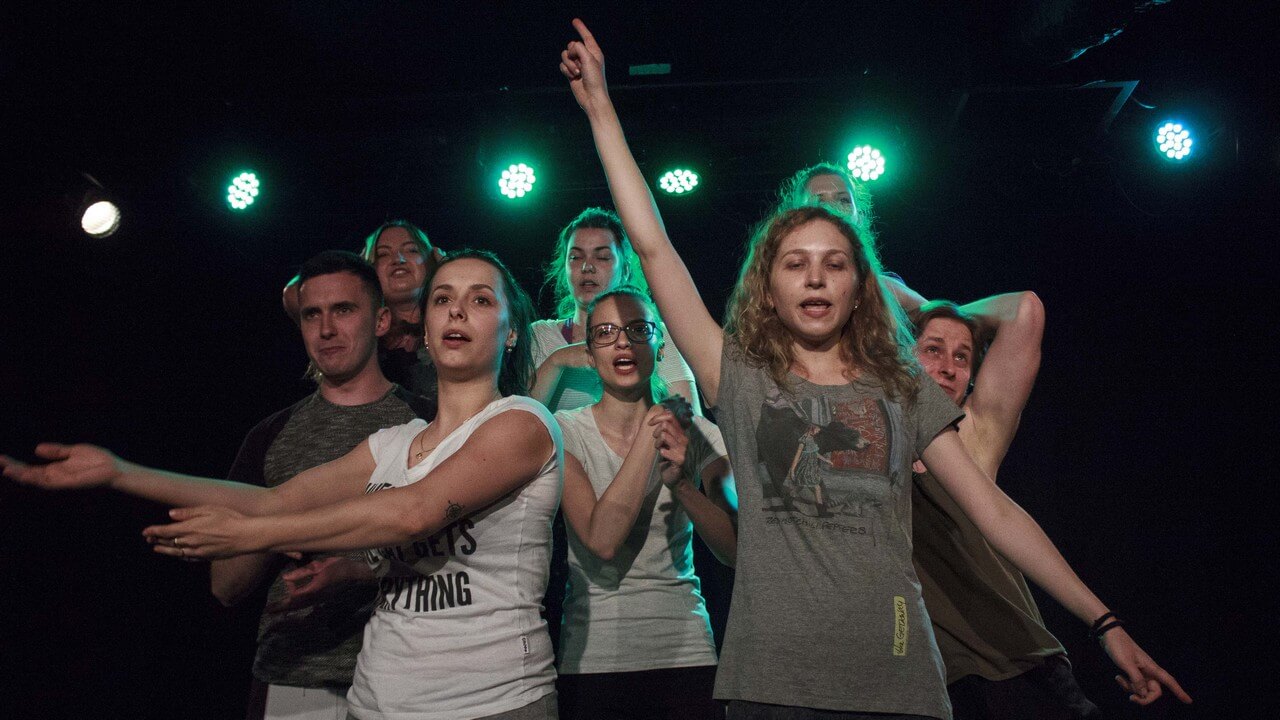 osiem osób stoi w grupie, wśród nich sześć kobiet i dwóch mężczyzn, wykonują dłońmi różne gesty, wyraz twarzy sugeruje, że coś mówią, kobieta na pierwszym planie wyciąga jedną rękę w górę, drugą w dół z tyłu pada zielone światło, zdjęcie z akcji w sieci teatru gdynia główna